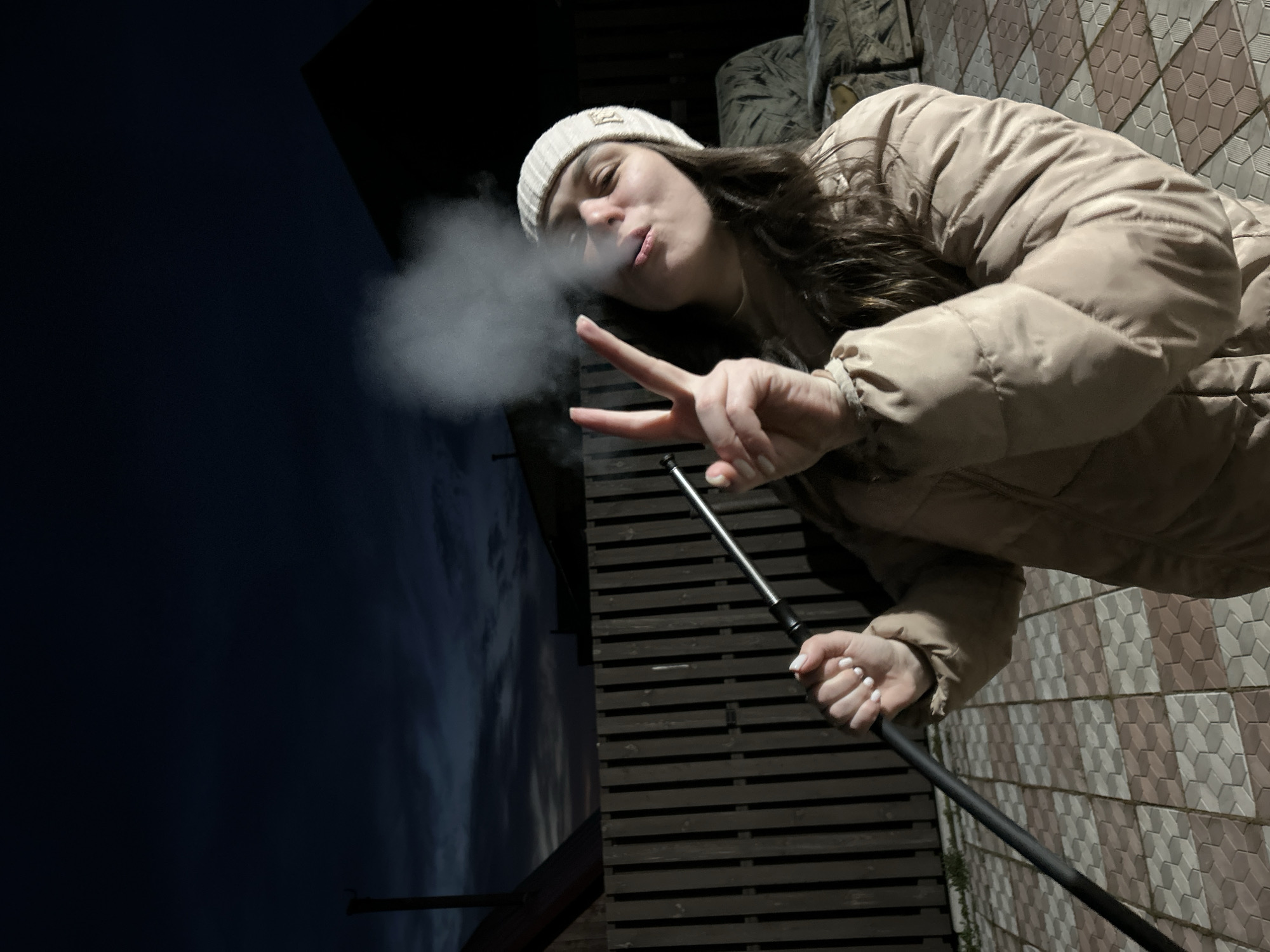 A woman with long brown hair wearing a cream-colored beanie and a light beige puffer jacket is exhaling smoke while holding a black hose. She is making a peace sign with her left hand. The background includes a dark night sky, the corner of a dark building, a wooden fence, and a tiled wall with a repeating geometric pattern. The location is Фоки, Russia.