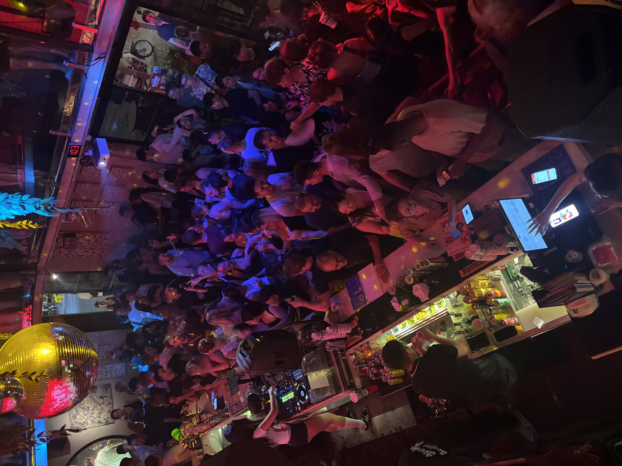 This photo shows a crowded bar or club in Đà Nẵng, Vietnam, seen from a slightly elevated perspective. A large group of people is clustered near the bar area, possibly waiting to order drinks. The lighting is colorful and dim, with a disco ball reflecting light onto the crowd. A DJ booth with equipment is visible near the front of the image, with a person standing and appearing to operate it. Behind the DJ booth, people are standing and dancing or socializing. The bar area has several employees serving drinks. The bar has alcohol bottles and glasses and is lit by LED screens. The room appears very crowded and energetic.