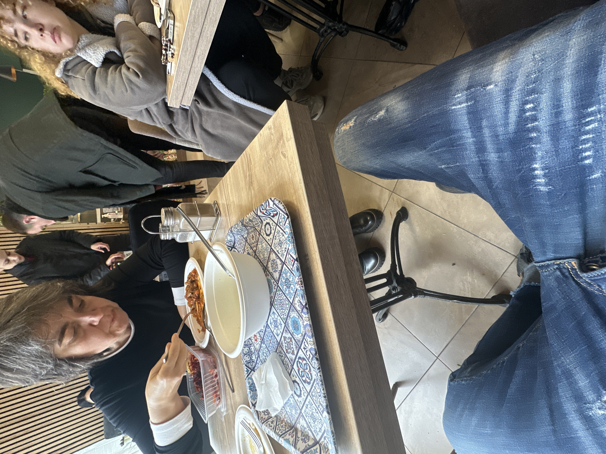 A tilted high-angle shot shows people dining in what appears to be a restaurant. Three women are visible, one with light-colored curly hair and wearing a brown coat, another with dark hair eating from bowls and containers on a patterned placemat, and a third partially seen. Additional people are blurred in the background. The environment includes a wooden table, visible parts of black chairs, and tiled flooring. A portion of a person wearing blue jeans fills the right side of the frame. The location is not specified but can be inferred to be somewhere in the city of Plovdiv, Bulgaria, assuming this information is provided.