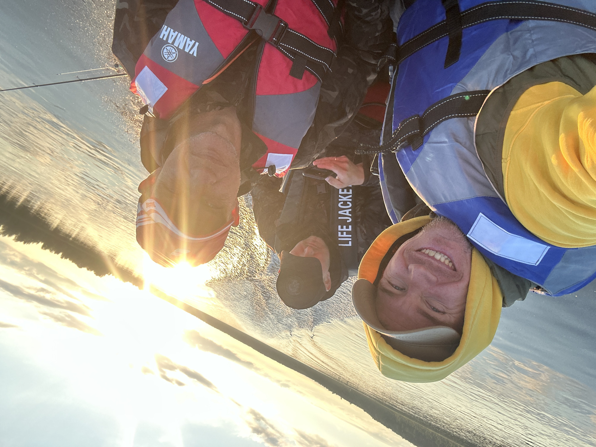 In this upside-down, wide-angle selfie, three men are captured from a low perspective on a body of water, likely a lake or river in Ol'khovskoye, Russia, given the location context. The setting appears to be a natural environment with calm water reflecting the sky and a distant tree line.
The subjects are wearing life jackets: one is red and grey with "YAMAHA" printed on it, another is blue and grey, and the third appears to be dark-colored. Two of the men are clearly visible in the foreground. The man on the right, wearing a yellow hoodie and a tan baseball cap, smiles broadly at the camera, his face illuminated by bright sunlight. His eyes are blue. The man in the center, wearing a dark baseball cap and a black jacket, also looks towards the camera, though his expression is less clear. The third man, partially visible on the left, wears a red life jacket and is looking slightly away from the camera.
The sun is low in the sky, casting a warm, golden light across the scene, suggesting it is either sunrise or sunset. The sky is partly cloudy, with some of the clouds illuminated by the sun. The water is disturbed, creating a rippled effect, which could indicate movement from a boat. There is a fishing rod visible in the upper left portion of the image.
A notable detail is the text "LIFE JACKE" visible on the black jacket of the man in the center, with the rest of the word obscured. The overall mood is jovial and relaxed, indicative of an enjoyable outdoor activity.