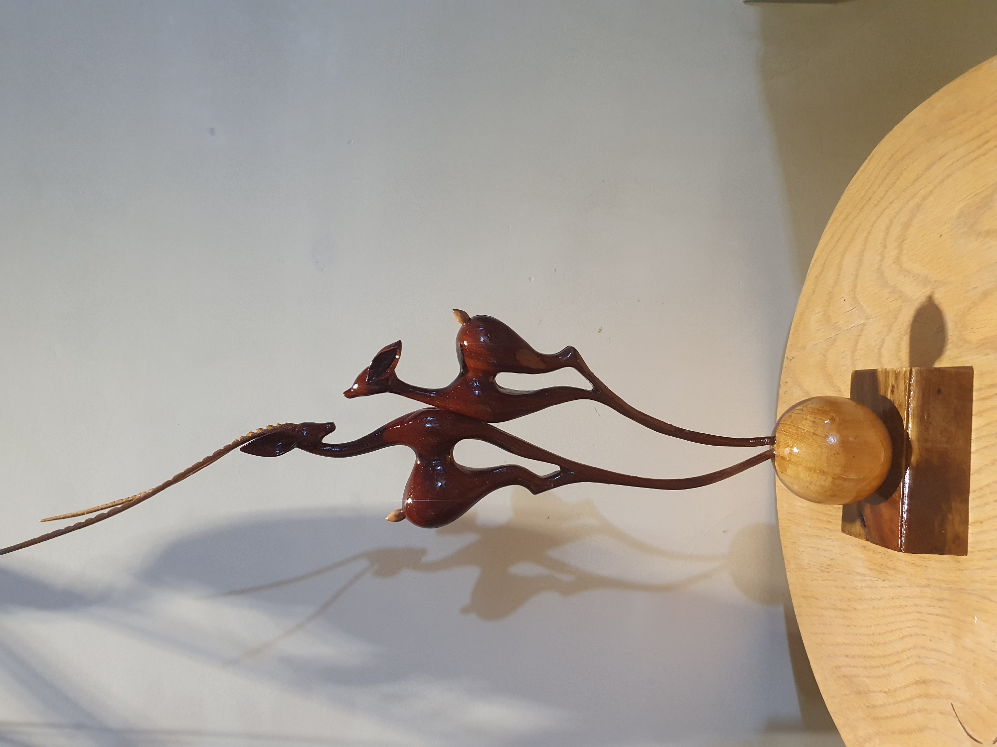 A wooden sculpture of two antelopes stands on a round, light-colored wooden table. The sculpture is dark reddish-brown and has a glossy finish. The antelopes are stylized, with slender bodies and long, thin legs. One antelope is slightly larger than the other and stands slightly behind it, facing to the left. The smaller antelope is positioned in front and to the right, facing left as well. Both antelopes have delicate, pointed ears and long, thin tails. A long, thin, twig-like element extends upwards from the head of the larger antelope, curving to the left. The sculpture is mounted on a base consisting of a small wooden sphere on top of a rectangular wooden block. The background is a plain, light-colored wall, with shadows cast by the sculpture. The lighting suggests an indoor setting, possibly a home or a shop. There are no people visible in the image. The scene is devoid of any explicit indicators of its location within the city of Shiraz, Iran.