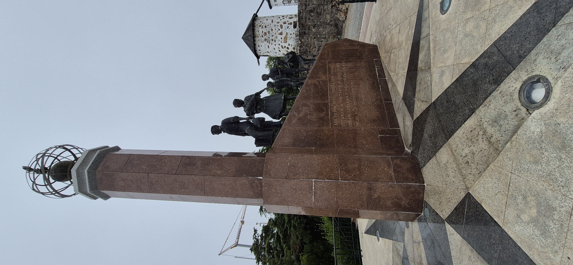 The image shows a monument with sculptures of people at its base, located in Póvoa de Varzim, Portugal. The monument is tall and rectangular, made of dark red granite, and topped with a globe-like structure. At the base of the monument, bronze sculptures depict a family, including a man, a woman holding a baby, and several children. The sculptures are positioned on steps leading up to the monument. An inscription is visible on the side of the monument. In the background, two old stone windmills with conical roofs are visible, along with lush green trees and a construction crane in the distance. The ground in front of the monument is paved with geometric patterns of light and dark stone tiles. The sky is overcast.