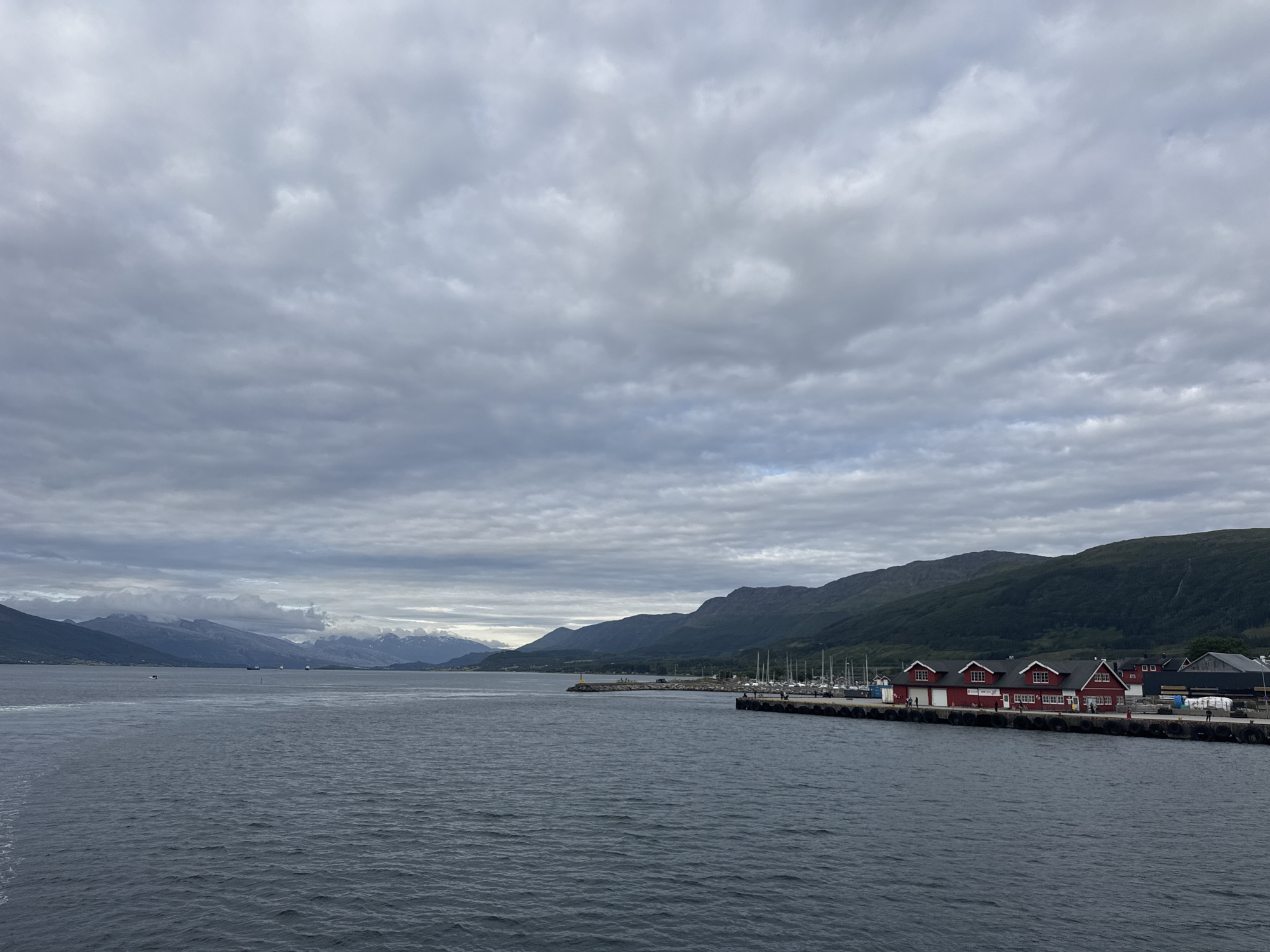 The image shows a waterfront scene in Nesna, Norway, under a cloudy sky. In the foreground, a dark, rippling body of water dominates the lower half of the frame. Along the right side of the image, a pier extends into the water, lined with several red buildings that appear to be boathouses or workshops, each with multiple dormer windows and dark roofs. A few figures are visible walking along the pier. Moored boats, primarily sailboats with tall masts, are docked behind the red buildings. Further out in the water, several larger ships are visible in the distance. The background features a range of mountains, with some peaks appearing snow-capped, and lush green slopes leading down to the water's edge. The overall atmosphere is overcast and somber.