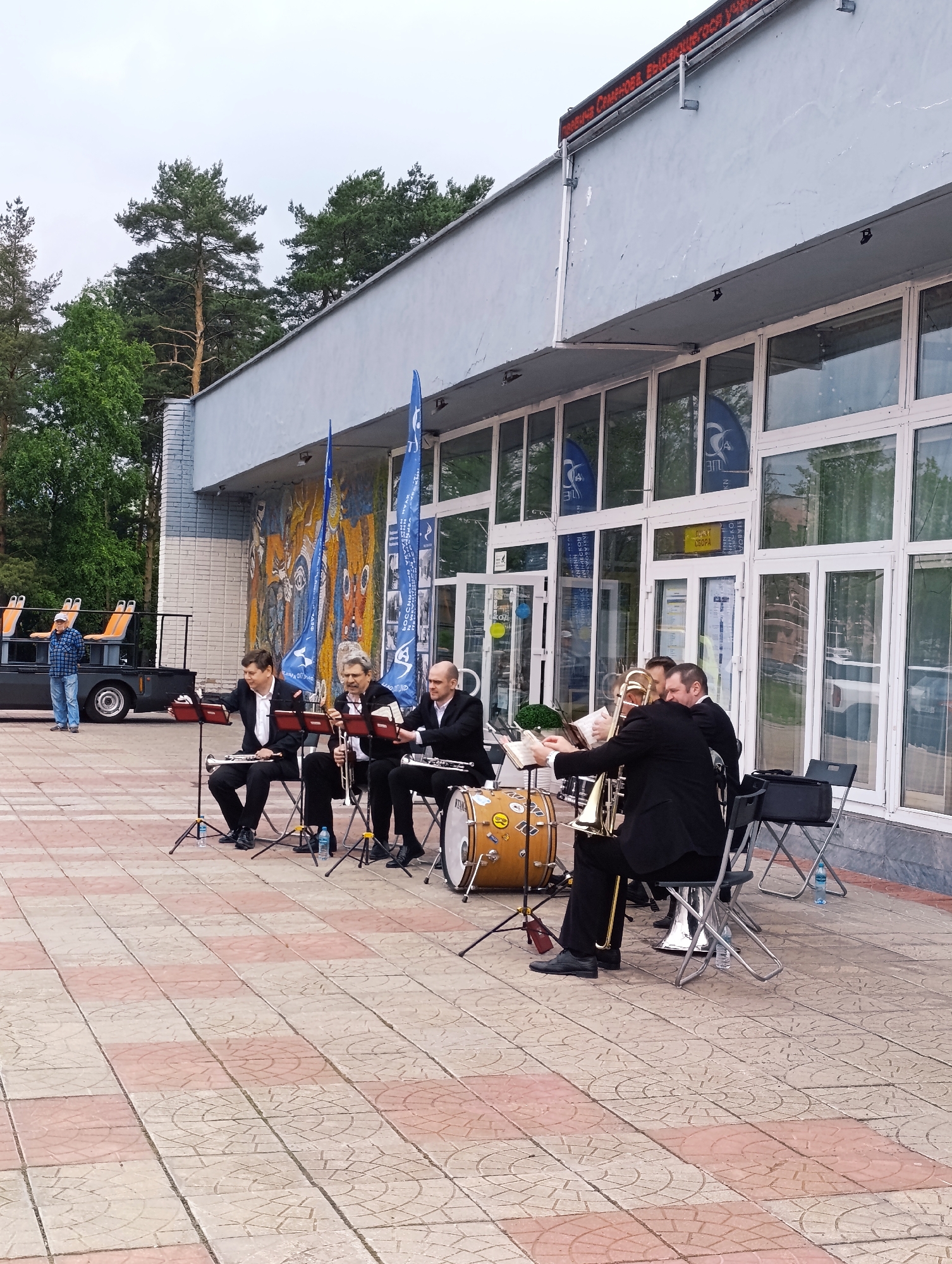 A brass band performs outdoors in front of a building with large windows and a mural on the wall. The band consists of five musicians in black suits playing trumpets, a trombone, and drums. To the left, a man in a blue plaid shirt and jeans stands near a utility vehicle with orange seats. The ground is paved with red and beige tiles. In the background, pine trees are visible. The sky is overcast. A digital sign with red text is mounted above the building's entrance.