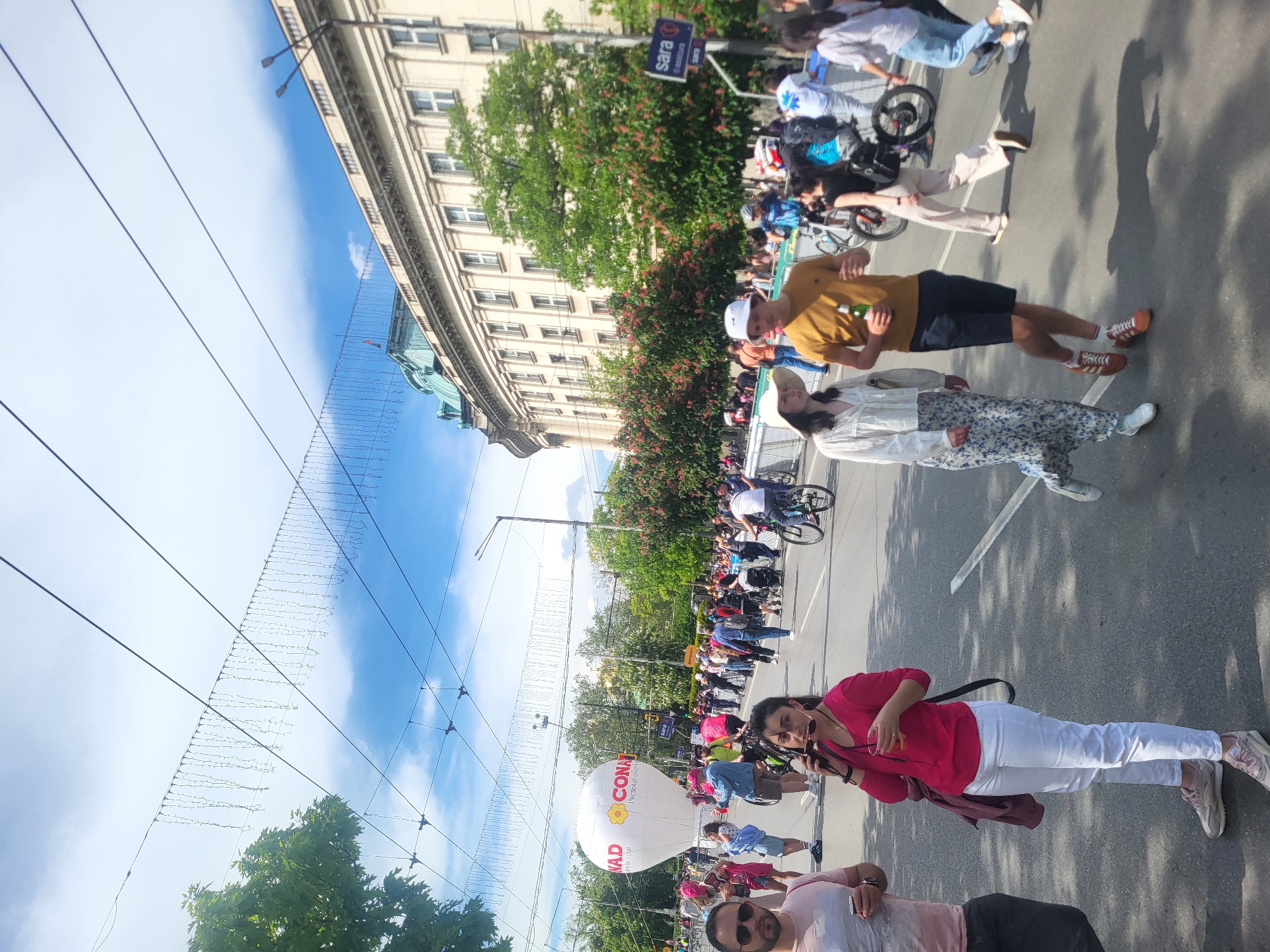 The image depicts a street scene in Unknown, Unknown, filled with a crowd of people. The crowd appears to be part of a parade or demonstration, as many are walking or cycling along the street. A woman in a pink shirt and white pants is prominently positioned near the bottom of the frame, holding what appears to be a camera. Behind her, a large white balloon with "CONAD" printed on it floats in the air. A sign with the text "sara" is visible on the right side of the frame. The background features a tall building, lush trees, and a blue sky with some clouds. Overhead wires with lights strung across them are also noticeable. The overall scene suggests a public event taking place in a city environment.