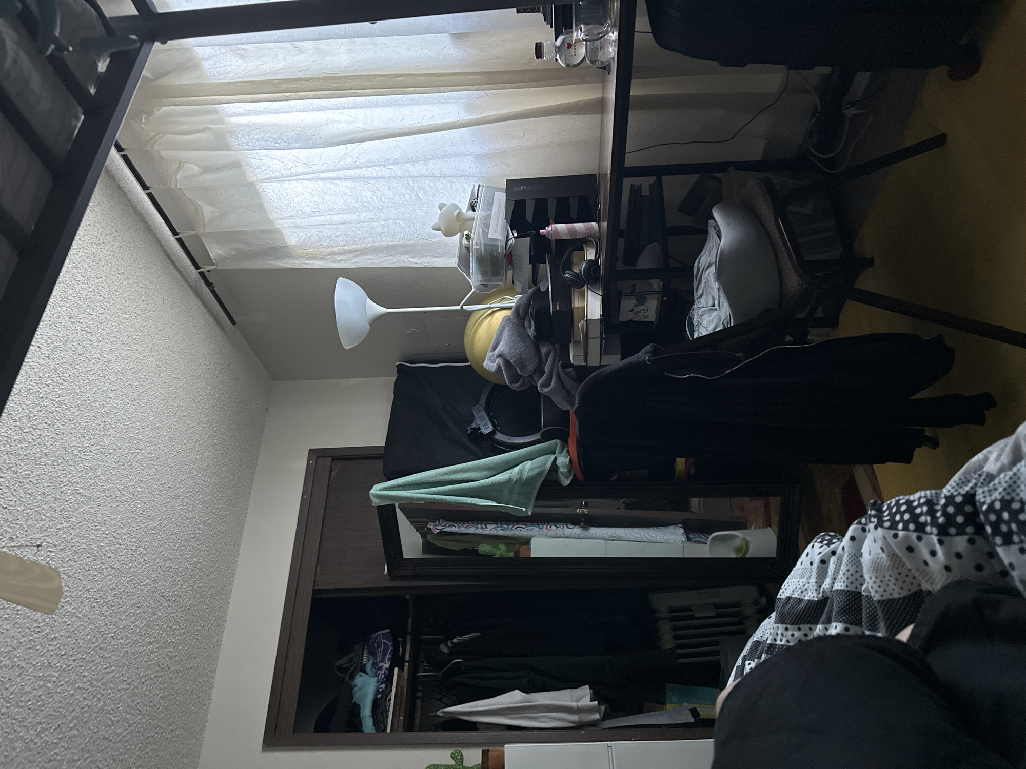 This image depicts an indoor scene, likely a bedroom, with a desk and a closet. The room is somewhat cluttered, with clothes hanging in the closet and various items on the desk. A floor lamp is positioned near the desk, and sheer curtains cover a window. A bed is partially visible in the foreground, covered with a patterned comforter. The overall impression is of a lived-in space. The image does not contain information that would allow for determining its location within the city of Vancouver, United States of America.