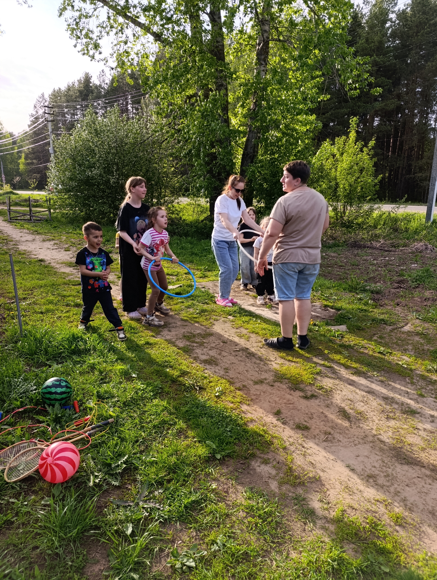 In Chaikovsky, Russia, a group of people stands on a dirt path surrounded by green grass and trees. There are at least five people visible, including adults and children. One child is holding a blue hula hoop. On the grass to the left, there is a red-and-white striped ball and two badminton rackets. In the background, there is a wooden fence and trees. The scene appears to be a casual gathering or outdoor activity.