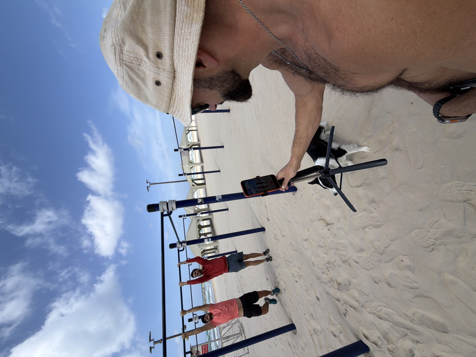 The scene is an outdoor calisthenics gym facility located on a sandy beach along the Tel-Aviv coastline, specifically characterized by multiple pull-up bar structures.
In the right foreground, a man, shirtless with a bearded face, wearing a beige bucket hat, dark sunglasses, and a silver chain, holds a smartphone in an orange case mounted on a black tripod. The phone is pointed towards the center of the frame, appearing to record or photograph the activity. A black and white dog stands on the sand near the base of the tripod, facing left.
In the mid-ground, two men are performing exercises on separate horizontal pull-up bars. The man on the left wears a coral t-shirt and dark shorts, hanging from the bar with both hands. The man on the right wears a red long-sleeved hoodie and dark shorts, also hanging from a bar with both hands. Both men have beards and are looking towards the camera/phone in the foreground.
The ground consists of light tan sand with visible footprints. In the background, the Mediterranean Sea is visible with gentle waves. Along the shoreline, multiple white-colored shaded structures, likely beach pergolas, are present. The sky above is clear blue with scattered white clouds. Tall light poles are visible in the far distance.