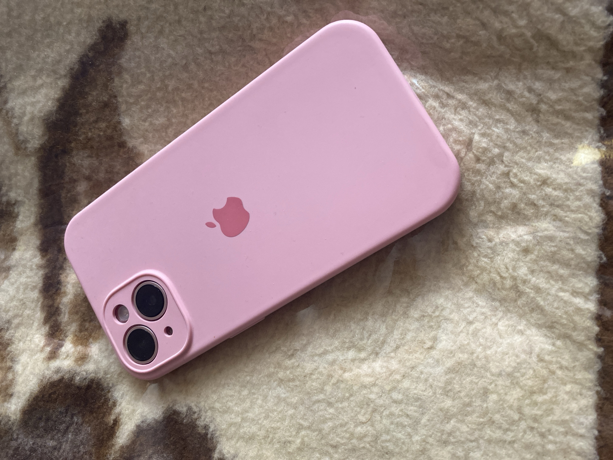 An iPhone 13 Pro in a pink case is shown from a top-down angle, lying on a textured surface. The surface appears to be a blanket or rug with a pattern of brown and cream-colored fibers. The iPhone's camera module, featuring two prominent lenses, is visible in the upper left corner of the device. The Apple logo is imprinted in a slightly darker shade of pink on the back of the phone. The lighting suggests an indoor environment, possibly a home. There are no people or other identifiable objects in the frame. The location of Chernogorsk, Russia is not discernible from the image.