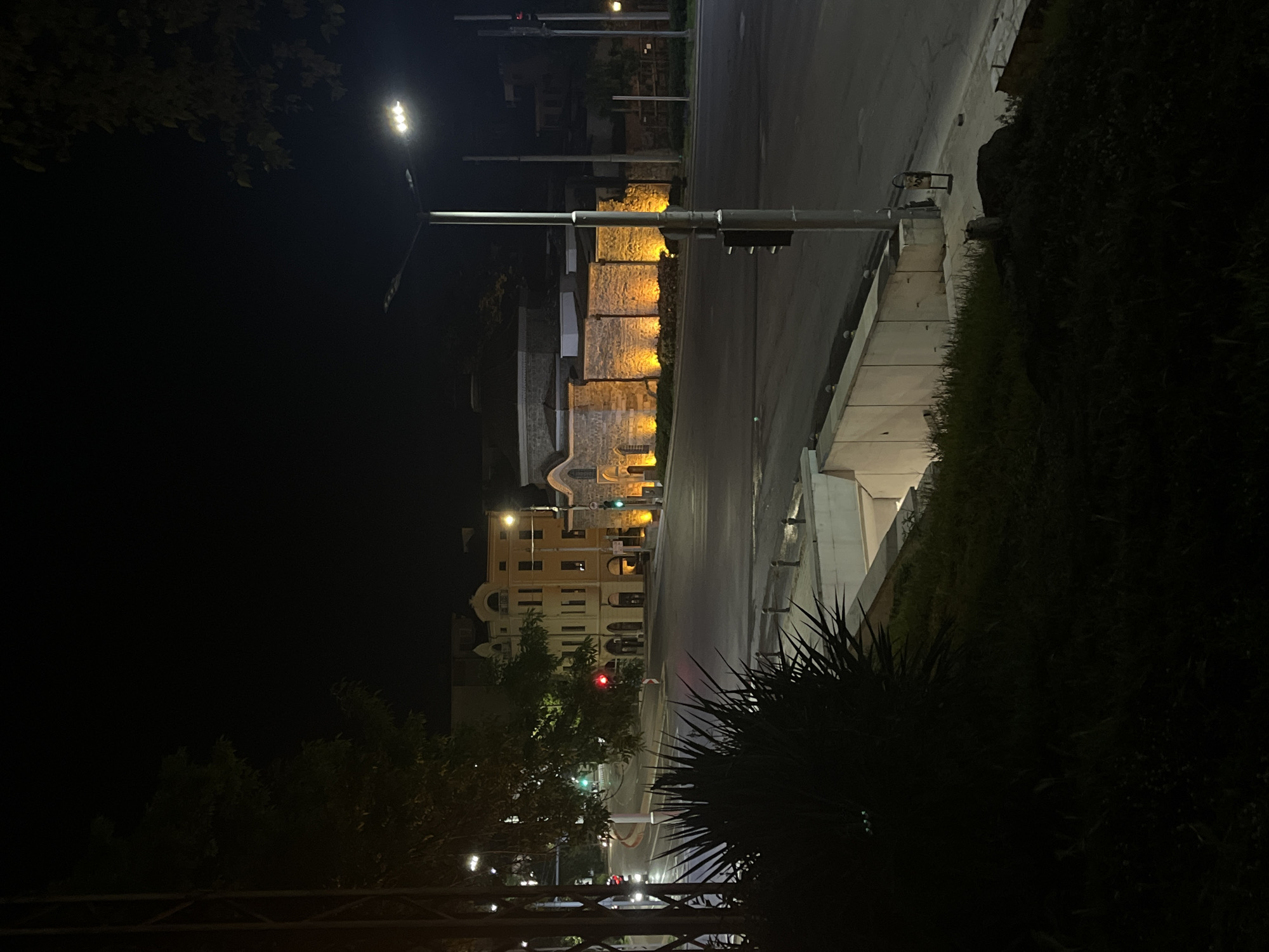 At night, a street view in Plovdiv, Bulgaria, shows buildings illuminated by streetlights. A road runs horizontally in the image, with light fixtures positioned along the left side. On the right, foliage obscures some of the view. Buildings are lit up, showing stone walls and windows. Traffic lights are visible. The image is taken at a slight angle.