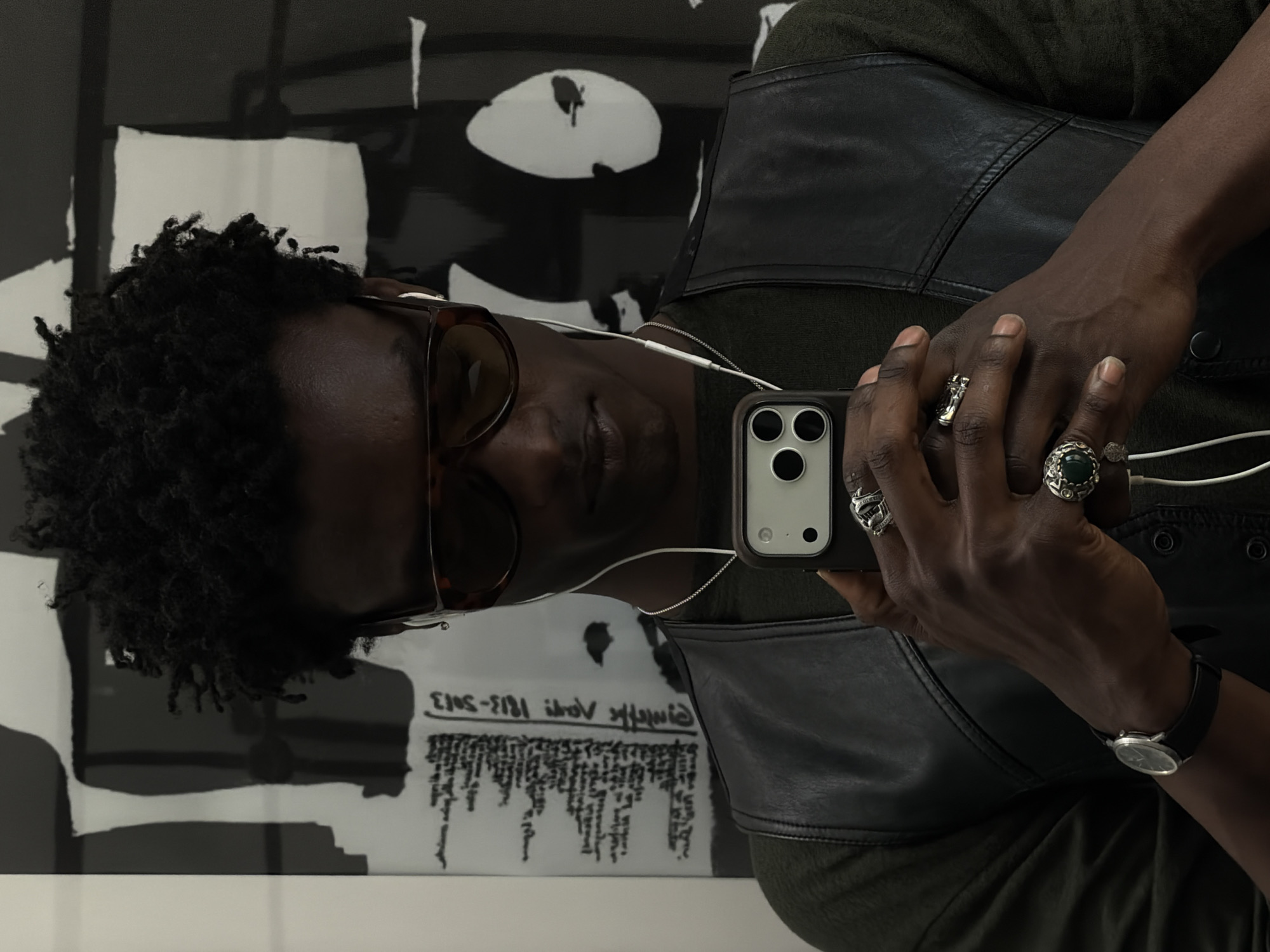 An indoor portrait depicts a person, likely in London, United Kingdom, holding an iPhone and appearing to interact with it, possibly taking a selfie or viewing the screen. The person has dark skin and short, dark curly hair, and is wearing tortoiseshell sunglasses. They are dressed in an olive green top with a black leather or faux-leather vest.
The person's right hand, which holds the phone, displays several rings: a prominent silver-toned ring featuring a large green stone, another silver-toned ring with an intricate crest or design, and a simpler silver-toned band. A silver-toned wristwatch with a black strap is visible on their left wrist. White wired earbuds are connected to the iPhone, which is in a light-colored case and clearly shows its triple-camera system.
The background features a large, abstract black and white artwork composed of bold, contrasting shapes. Reflected on the surface of this artwork, reversed, is text that reads "Giafalle 1813 - 3013," along with a block of smaller, illegible text below it. The lighting is bright and consistent, suggesting an indoor setting such as an art gallery or exhibition space. The sunglasses obscure the person's expression, making their emotions difficult to discern.