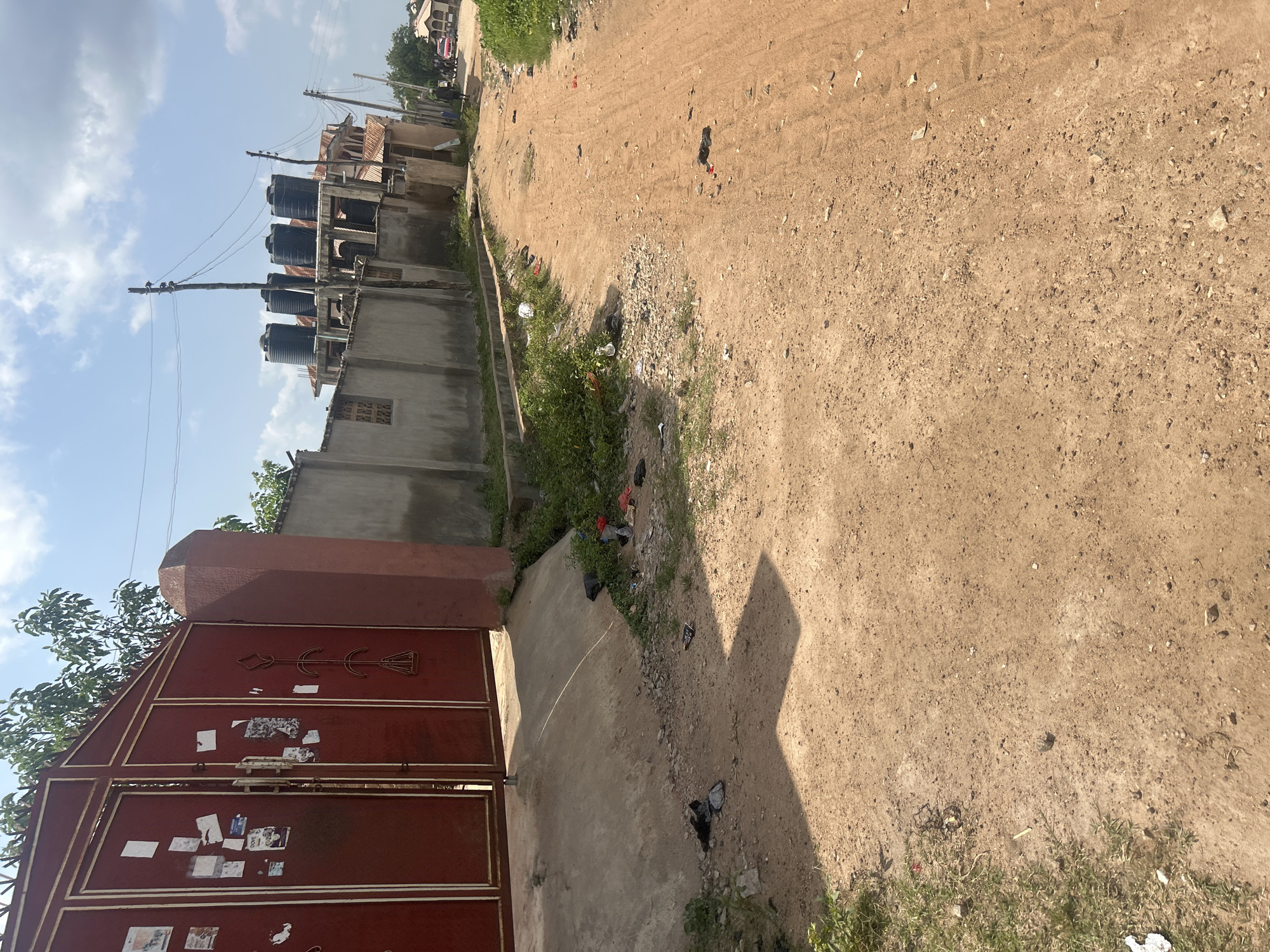 The image depicts a street scene in Ogbomosho, Nigeria. The foreground features a large, closed, maroon-colored metal gate with decorative elements and several pieces of paper affixed to its surface. To the left of the gate, a concrete pathway leads away from the viewer. Beyond the gate and pathway, a concrete wall runs along the property line.
Dominating the mid-ground on the left are several large black water tanks mounted on elevated structures, likely for water storage. These structures appear to be part of multi-story buildings with corrugated metal roofs. Electrical poles and wires are visible against a bright sky. The right side of the image shows a dirt road with scattered debris and patches of dry grass. In the distance, more buildings with similar roofing can be seen. The overall impression is that of a residential or semi-industrial area under a clear, sunny sky, suggesting it is daytime. There are no people actively engaged in any visible activity, and the scene is static, conveying a sense of everyday life in this location. The presence of the water tanks suggests a potential concern with water supply, common in some regions. The paper on the gate is too small to be legible.