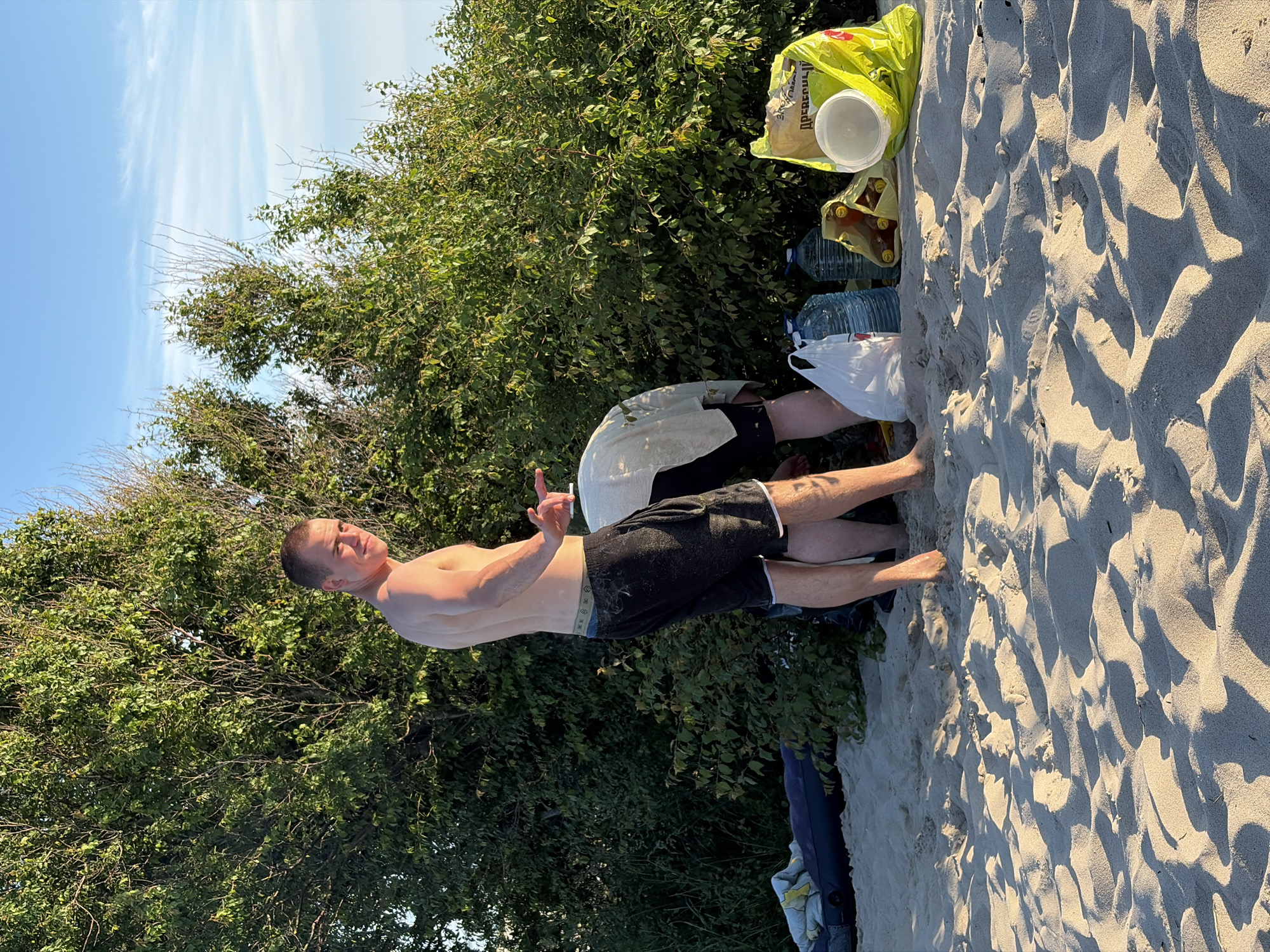 A young man stands on a sandy beach in Povarovka, Russia, facing away from the camera and looking towards a dense green bush. He is shirtless and wearing black shorts. Another person, also shirtless and wearing a light-colored garment over their lower body, is crouched down near the bush, seemingly engaged in an activity. In the foreground, the sand is textured with shadows from the setting sun, and various items are scattered on the beach, including plastic bags, large water bottles, and a yellow bag with a white bowl inside. The sky above is a clear blue with a few wispy clouds.