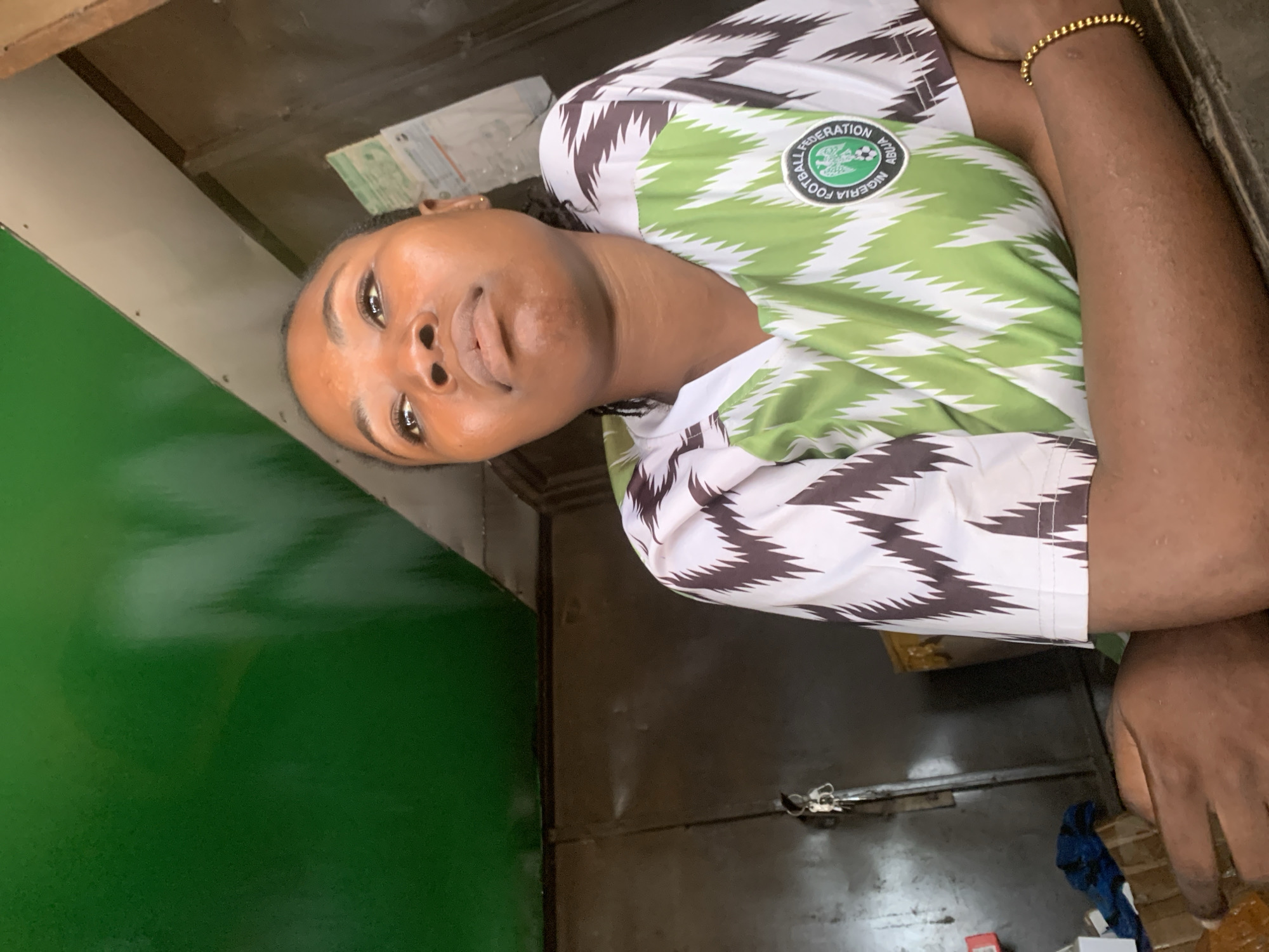 A young Black woman, with dark hair pulled back and dark eyes, is pictured from the chest up, looking upwards and to the right. She is wearing a green and white patterned jersey with a circular emblem on the left side of her chest that reads "NIGERIA FOOTBALL FEDERATION ABUJA". Her arms are crossed, and she has a gold beaded bracelet on her right wrist. She is seated in what appears to be an indoor market stall with a green ceiling and dark, utilitarian walls. The background shows some shelves and hanging items, suggesting a commercial environment. This scene is set in the city of Shagamu, Nigeria.