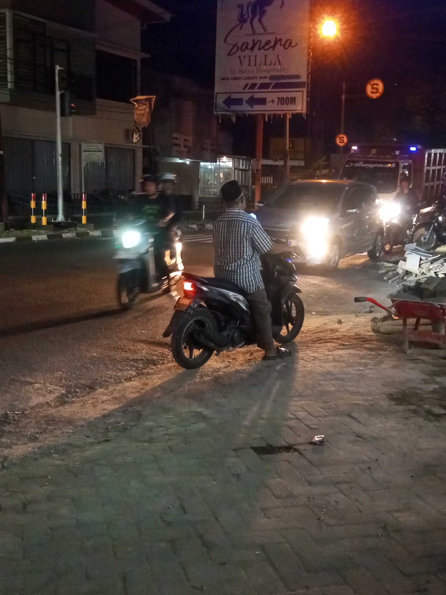 This is a nighttime photograph taken on a street in Wonosobo, Indonesia. The scene depicts a busy street with several vehicles and pedestrians. In the foreground, a man wearing a checkered shirt and a black cap is seated on a scooter, facing away from the camera. His scooter is parked on the side of the road, near a pile of construction debris that includes a red wheelbarrow and various building materials.
Further down the street, another scooter with a rider and a passenger is moving away from the camera, its headlight illuminated. To the left, a traffic light is visible on a pole, with a banner hanging from it.
In the background, a dark-colored car with its headlights on is visible, along with a truck with "Anugrah" written on its side, illuminated by blue flashing lights. A street sign with the text "Sanera VILLA" and "INFO LEBIH LANJUT HUB -- 700M" along with arrows pointing left is also present, alongside a circular sign with the letter "S" in orange. The street is paved with interlocking bricks, and the overall lighting suggests it is well-lit by streetlights and vehicle headlights. The environment appears to be a commercial or residential area with buildings visible on the left side of the street.