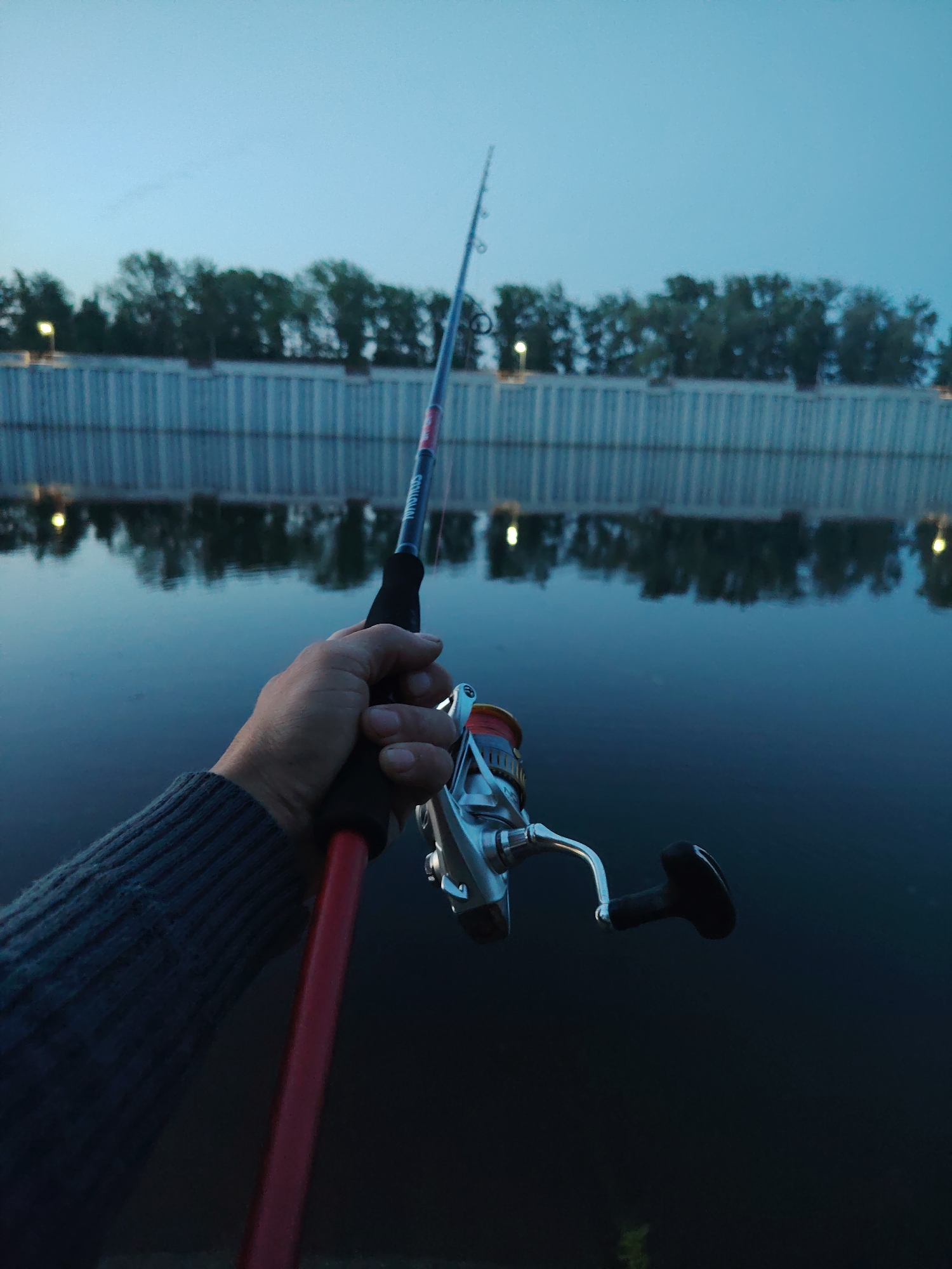 A person holds a fishing rod with a reel attached in front of a body of water. The water is dark and reflects lights and trees along the far bank, which is lined with a light-colored concrete barrier. The sky is a gradient of blue, suggesting either dusk or dawn. The person's hand is gripping the rod's handle, and their arm, clad in a gray knitted sweater, is visible. Based on the architecture of the embankment, the scene may be the city of Chaikovsky, Russia.