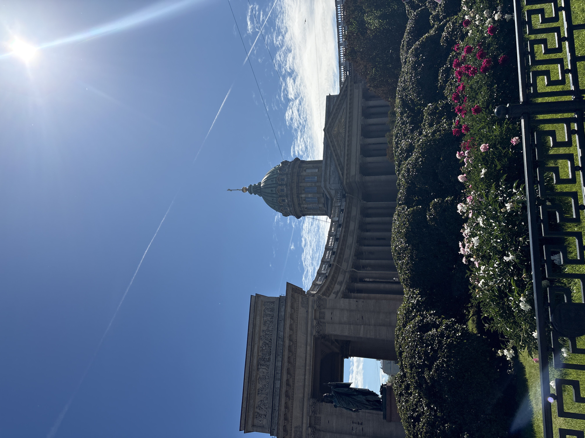 The image shows a view of Kazan Cathedral in Saint Petersburg, Russia, under a bright, sunny sky with contrails from airplanes. In the foreground, a section of a black metal fence with a geometric pattern is visible. Behind the fence, a manicured lawn is present, and further back, lush green bushes are densely planted. Blooming flowers, primarily white and pink peonies, add vibrant color to the scene in front of the bushes.
To the left, a portion of the cathedral's colonnade is seen, with a large, dark bronze statue of a figure standing in front of an arched entryway. The colonnade curves around to the right, leading towards the main body of the cathedral, which features a large, green dome topped with a cross. The architecture is classical, characterized by numerous columns and detailed ornamentation. The bright sun creates a lens flare effect in the upper right portion of the image. The overall atmosphere is one of grandeur and clear weather.