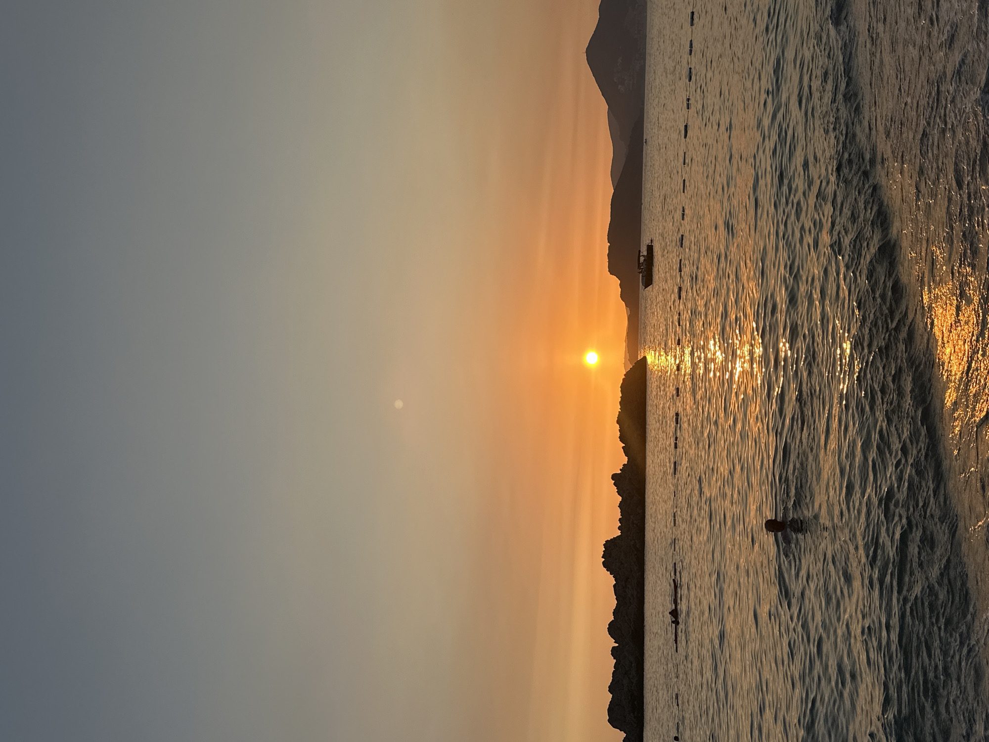 This image captures a serene sunset over the Adriatic Sea, with the iconic island of Sveti Stefan in Montenegro visible in the distance. The sun, a vibrant orb of orange, is low on the horizon, casting a warm, golden glow across the sky and water. Its reflection shimmers on the rippling surface of the sea, creating a path of light that leads towards the shore.
In the foreground, a person is swimming in the calm waters, their head and shoulders visible above the surface. The water is a deep blue-green, with gentle waves lapping at the sandy shore. In the mid-ground, a rocky outcrop juts out from the sea, its dark silhouette contrasting with the bright sky. A small boat, silhouetted against the sunset, cruises in the distance near the base of the island. The island itself, Sveti Stefan, is a fortified medieval village perched on a rocky islet, connected to the mainland by a narrow causeway. The buildings are rendered in silhouette, their red-tiled roofs a familiar sight.
The sky transitions from a soft blue at the top to a warm orange and yellow near the horizon, painted with the subtle hues of a setting sun. The overall atmosphere is one of tranquility and natural beauty, a perfect depiction of a summer evening in Montenegro.