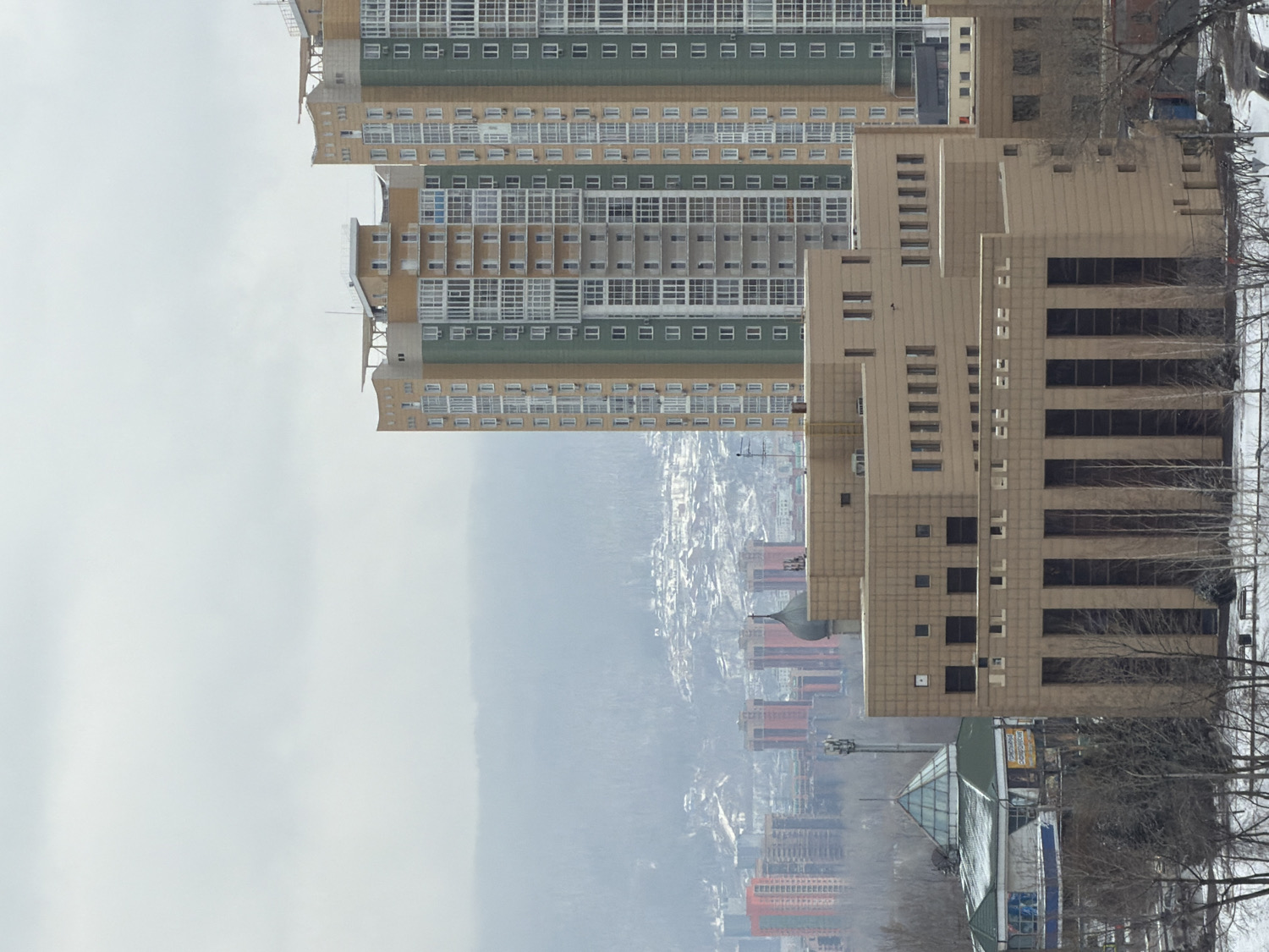 The image displays an urban landscape, rotated 90 degrees clockwise from its correct upright orientation, featuring several tall buildings and a distant city sprawling across a hazy valley under an overcast sky. The setting is Krasnoyarsk, Russia.
In the foreground, on the right side of the frame (when viewed correctly), stands a prominent multi-story building of light beige or tan stone, characterized by a stepped, terraced design with numerous rectangular and square windows. Adjacent and behind it are multiple large residential apartment blocks, distinguished by horizontal bands of alternating light beige/tan and dark green/grey facades, with many grid-patterned windows.
The midground and background reveal a vast urban expanse, with more buildings of varying sizes extending into the distance. The distant landscape appears hilly or mountainous, covered in a dense network of structures that become increasingly obscured by atmospheric haze or fog. The sky above is entirely overcast, suggesting a cloudy day with diffuse lighting.
On the far right (when upright), and in the lower right corner, several bare deciduous trees with dark, slender branches are visible, indicating either winter or early spring. Patches of snow can be seen on the ground near these trees. Below the prominent beige building, closer to the bottom of the frame (when upright), a structure with a glass roof is partially visible.
No people or explicit activities are depicted, giving the scene a quiet, somewhat stark atmosphere typical of a cold, overcast day.
**Visible Text:** On a building facade in the lower right portion of the image (when upright), a yellow sign is visible with Russian text that reads "СПОРТМАСТЕР" (SPORTMASTER), indicating a sporting goods store.