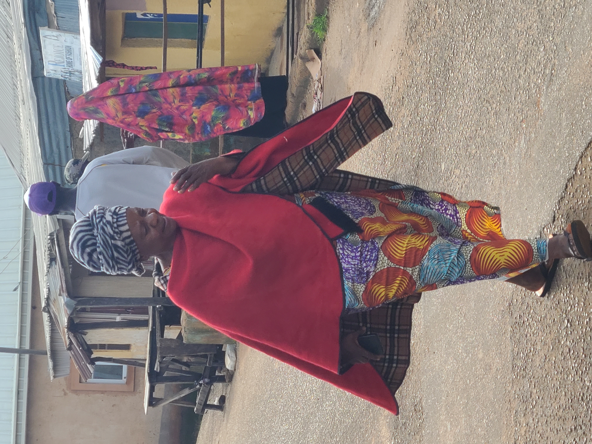 A woman is walking on a dirt road in Jos, Nigeria. She is wearing a red shawl over a colorful patterned dress and a black and white striped headwrap. Her right hand is extended forward, and she appears to be holding a mobile phone. In the background, there are several buildings with corrugated metal roofs, and a sign that reads "REJOICE HOME OF FASHION." Another person, wearing a purple baseball cap and a white shirt, is visible further down the road, as is a person in a brightly colored headscarf. The ground is paved with asphalt, and there is some debris scattered on the roadside. The lighting suggests it is daytime, with the sun casting shadows.
