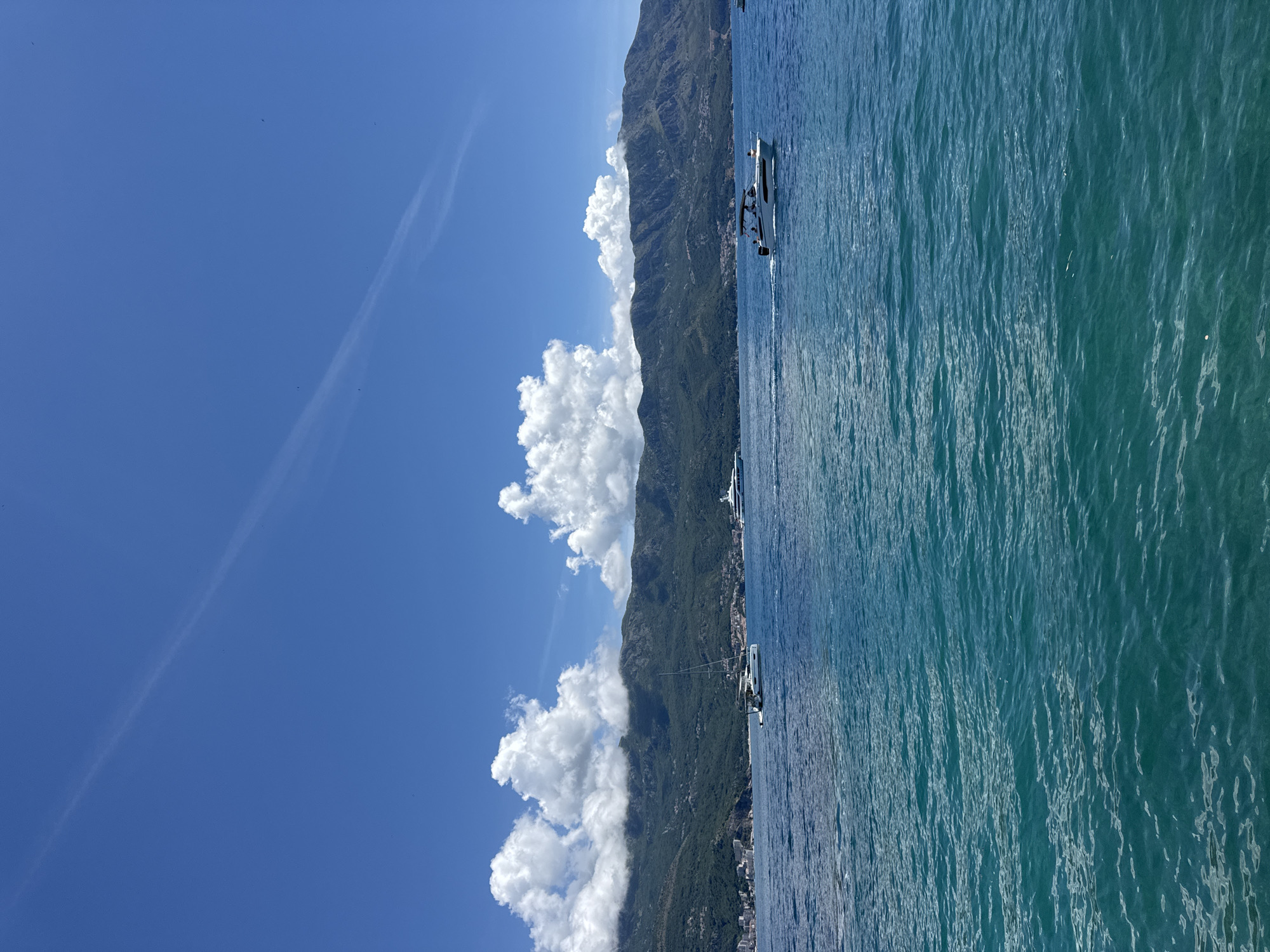 An aerial or elevated view captures a serene coastal landscape in Budva, Montenegro, under a bright blue sky. The image is dominated by the expansive, clear blue water of what appears to be the Adriatic Sea on the right, showing ripples and variations in blue and green hues. On the left, a majestic mountain range rises, densely covered in vibrant green foliage. White, fluffy cumulus clouds are scattered across the sky, contrasting with the deep blue. A faint white contrail streaks across the upper left sky, with a few tiny dark specks potentially being birds.
Several white motorboats or yachts are visible on the water near the mountainous coastline, suggesting recreational activities. One boat in the mid-ground appears to have a few indistinct figures on board. In the distance, at the base of the mountains, faint architectural structures hint at a coastal settlement. The scene depicts a clear and sunny day, likely midday or early afternoon, with ideal weather conditions for boating and outdoor enjoyment. The overall mood is one of peace and natural beauty. No discernible text is visible in the image.