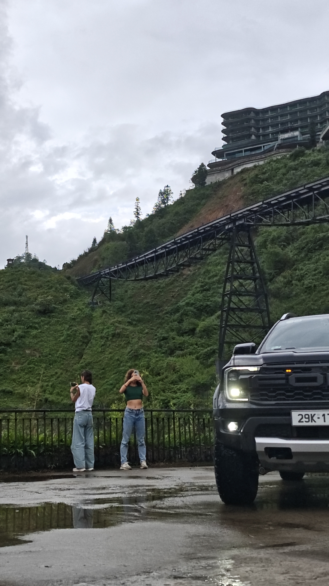 Two women stand on a paved surface, holding their phones as if taking pictures. One woman wears a white shirt and blue jeans, while the other wears a green crop top and blue jeans. A black Ford pickup truck is parked to the right of the frame. Behind the women, a metal bridge spans a green, hilly landscape, with a building visible atop the highest point. The sky is overcast. The visible license plate on the truck reads "29K-17". The location appears to be near Cat Cat Village, Vietnam. The ground is wet, and reflections are visible.