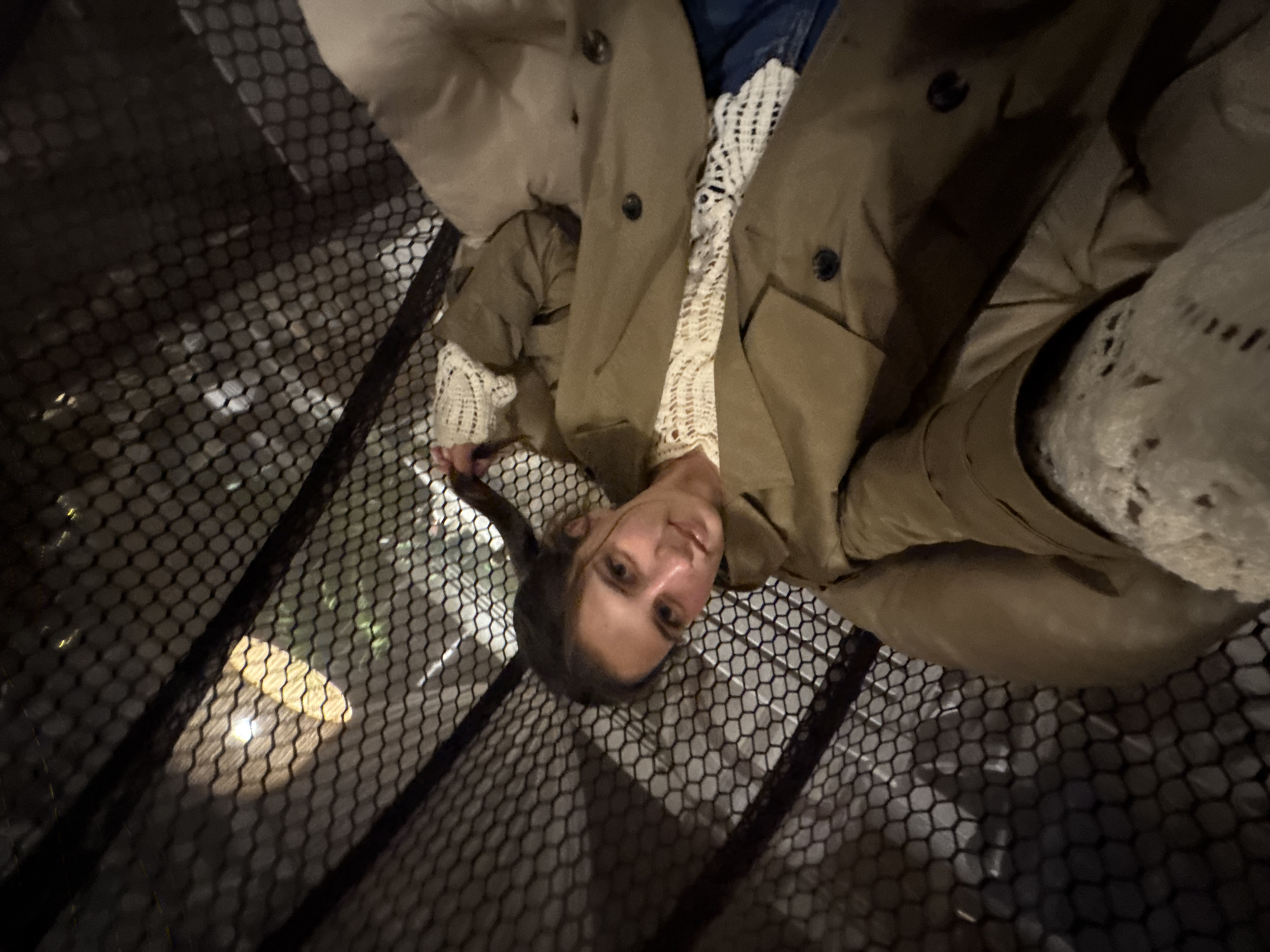 A young woman is captured in an upward-looking shot, lying on her back on a dark, hexagonal mesh surface. She gazes directly at the camera with a neutral to slightly smiling expression. Her dark hair is styled back, revealing her face. She is dressed in a light-colored, possibly crocheted or knitted top, worn underneath a functional-style olive-green or khaki trench coat or jacket. Her upper body is visible, including her arms and the folds of her coat.
The prominent black mesh forms a patterned foreground and midground, suggesting she is on a suspended net or a mesh floor within a multi-level structure. Below the mesh, blurred lights are visible, indicating an illuminated space below, possibly another floor or the ground level. The overall lighting is dim, suggesting an indoor setting during the evening or nighttime. The unique perspective, looking down through the mesh, gives the photograph an unusual and dynamic quality. There is no visible text in the image. The scene is located in Perm, Russia.