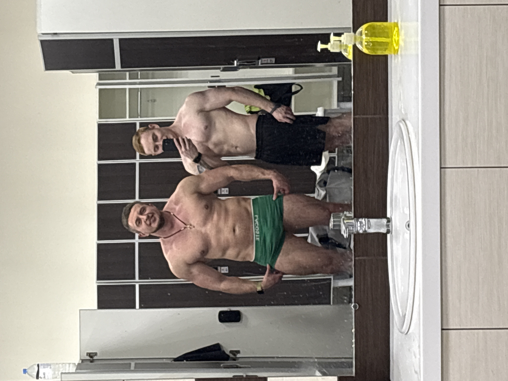 Two men are reflected in a mirror in a bathroom or locker room setting. The man closer to the mirror is muscular and is wearing green underwear with "LACOSTE" visible. The second man, positioned behind and slightly above the first, is holding a phone and appears to be taking the picture. He is wearing black shorts. A sink, soap dispenser, and a water bottle on the counter are visible in the foreground. The background shows lockers. There is no verifiable evidence of the city location.