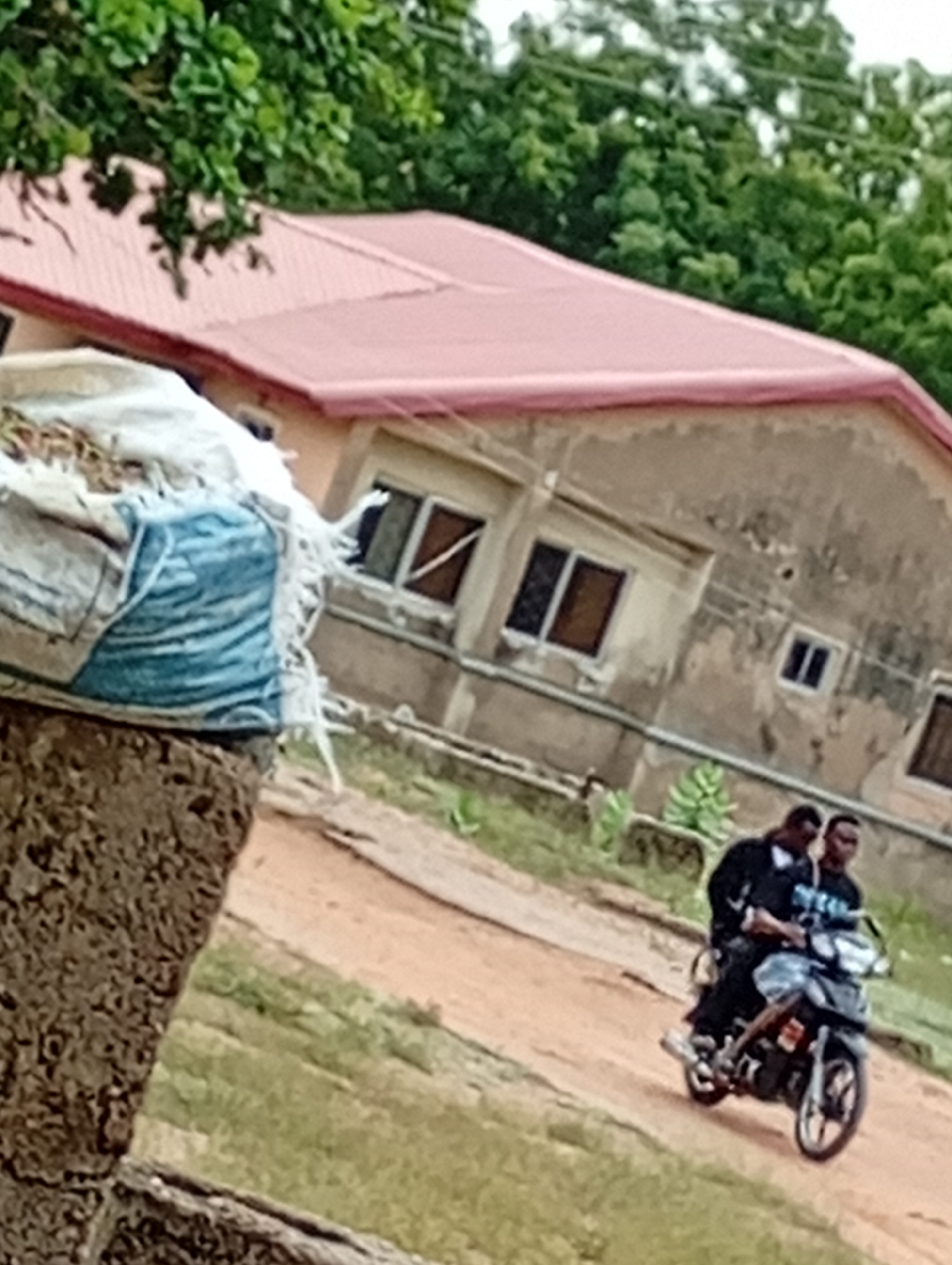 The image shows two people riding a motorcycle on a dirt road. In the background is a building with a red roof and several windows. The location appears to be a rural or semi-rural area. The specific city and country are unknown. In the foreground, there is a concrete structure with a blue and white bag on top.