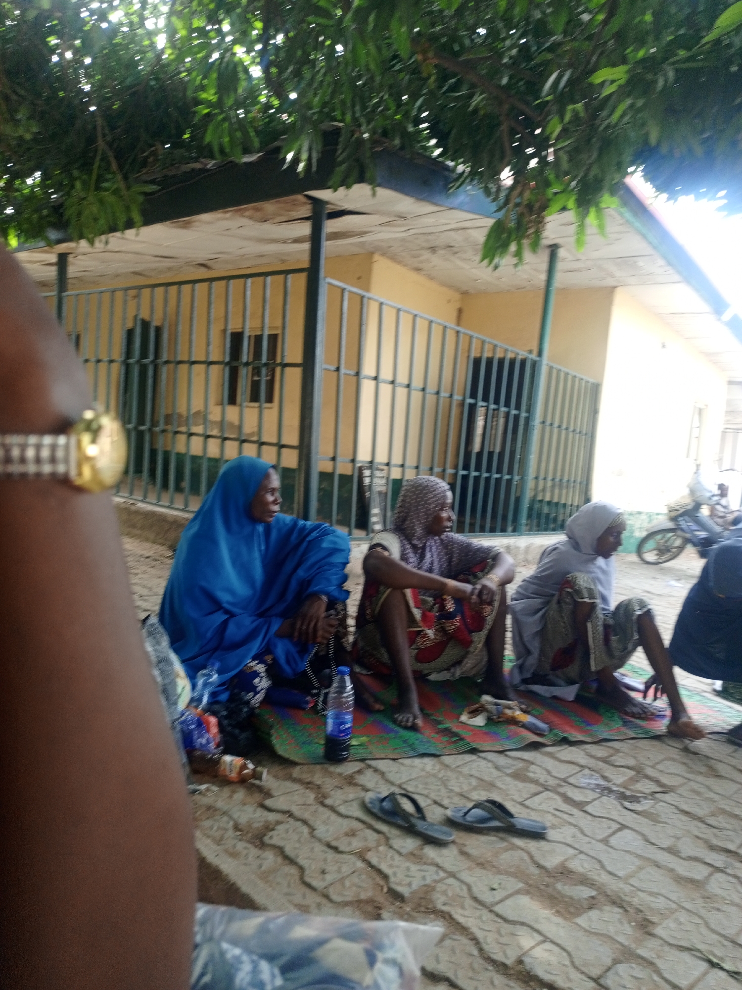 Three women in traditional Islamic clothing sit on a mat on a paved surface, with a building featuring barred windows in the background. The scene is shaded by foliage. Drinks, shoes, and bags are scattered around the mat. A motorcycle is parked nearby. The location is identified as Keffi, Nigeria.