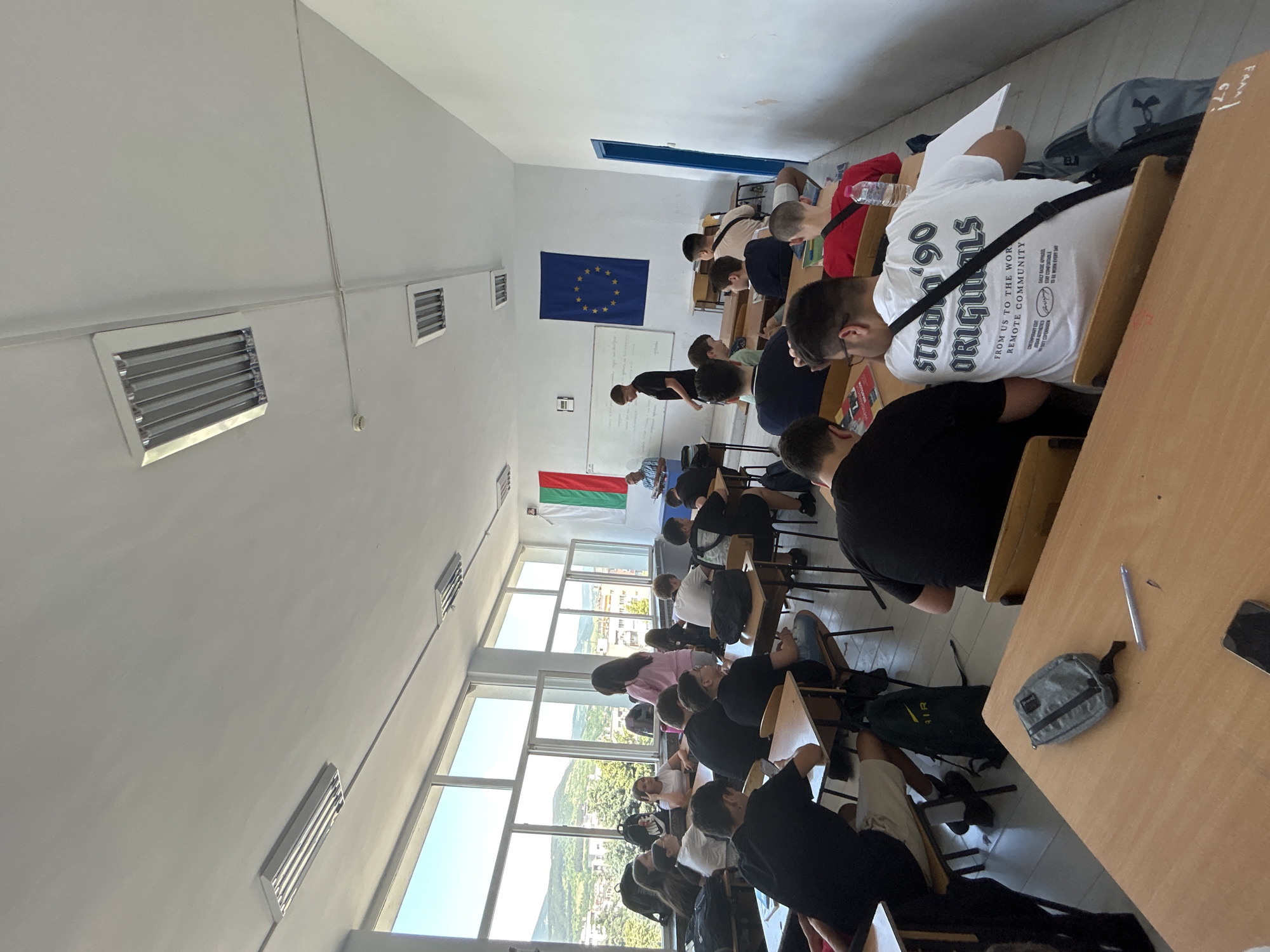 The image shows a classroom setting, presumably in a school in Kardzhali, Bulgaria. Students are seated at desks, many appearing to be taking a test or working on an assignment. The room has rows of desks facing towards the front, where there's a whiteboard. The Bulgarian and European Union flags are hung on the wall. Windows line one side of the room, offering a view of the city and surrounding landscape. The lighting is bright, and the students are dressed in casual attire. There are backpacks and personal items near the desks. The image suggests an educational environment where a test or assignment is in progress.