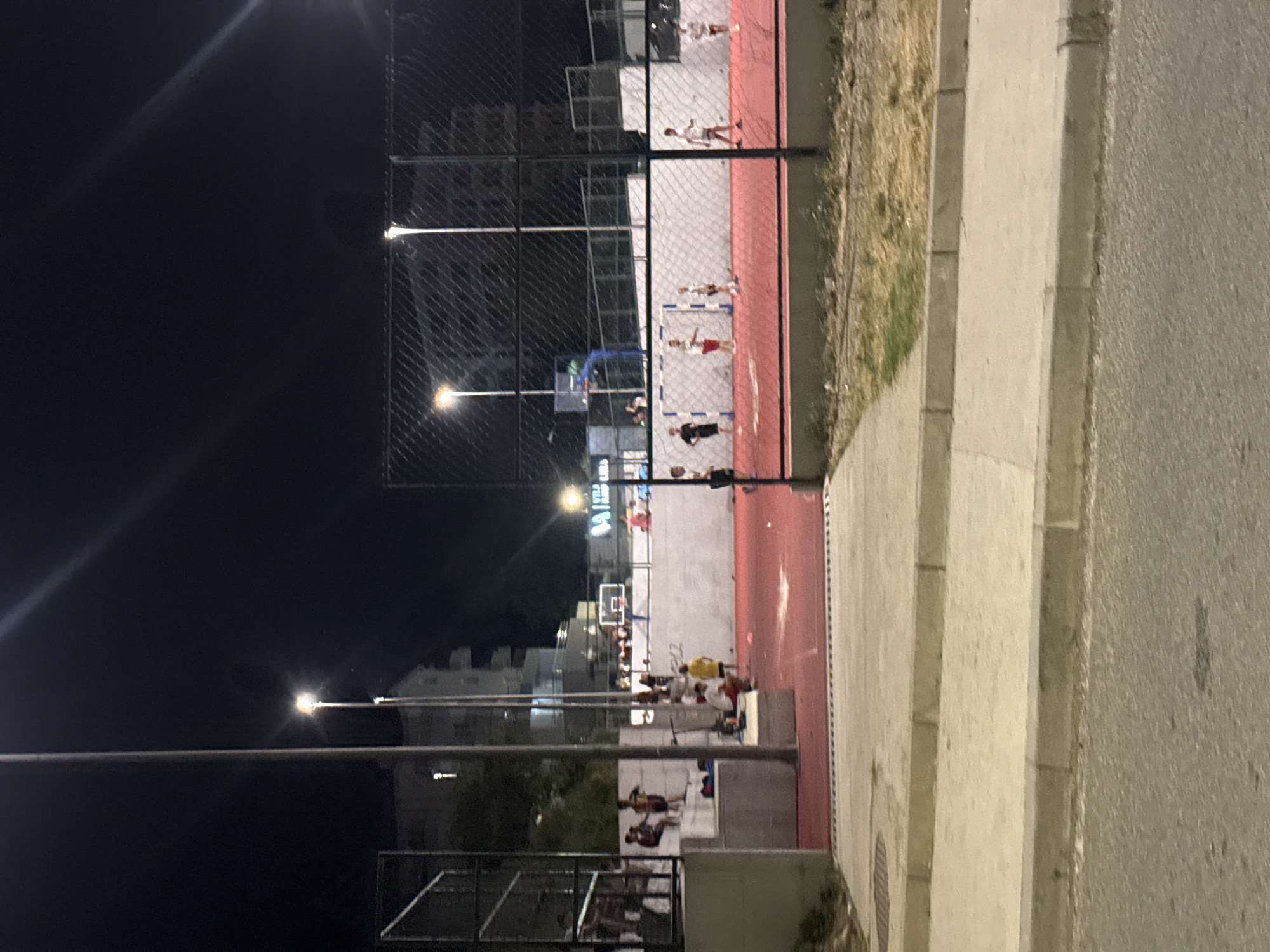 The image depicts an outdoor sports court illuminated by artificial lighting at night in Boreti, Montenegro. The court has a red playing surface and is surrounded by a fence. Several individuals, appearing to be children and young adults, are present on and around the court. Some are actively engaged in sports, such as playing with a ball on the court, while others are standing or sitting on elevated concrete structures. In the background, buildings and trees are visible, along with streetlights. The scene suggests a public recreational area being utilized in the evening.