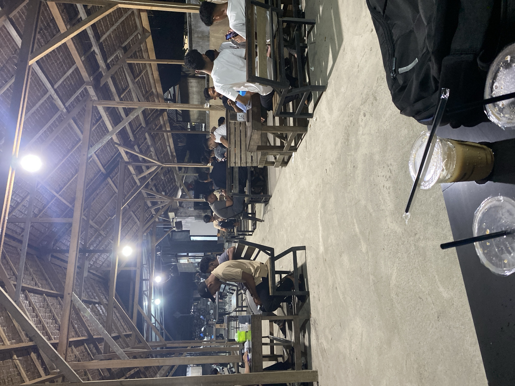The image captures an indoor or semi-outdoor gathering place at night in Seuneubok Teungoh, Indonesia. The scene is illuminated by several bright lights, suggesting evening hours. The structure features a distinctive wooden beam framework and a thatched roof, giving it a rustic or traditional aesthetic.
Numerous individuals, predominantly men, are seated at wooden tables and chairs arranged in rows. Many appear to be engaged with their mobile phones. One person in the foreground, wearing a light-colored shirt and a dark baseball cap, is leaning forward on the table.
In the immediate foreground, on a dark table, there is a black padded backpack. Beside it, three plastic cups are visible; one contains a light brown beverage, possibly iced coffee, with a straw, while the other two appear mostly empty with straws. The table surface is a concrete or light-colored floor.
A notable detail is a white card with the number "16" printed on it, visible on one of the wooden tables in the midground. The overall atmosphere appears casual and social. The image is oriented vertically, rotated 90 degrees clockwise from its natural horizontal perspective.
