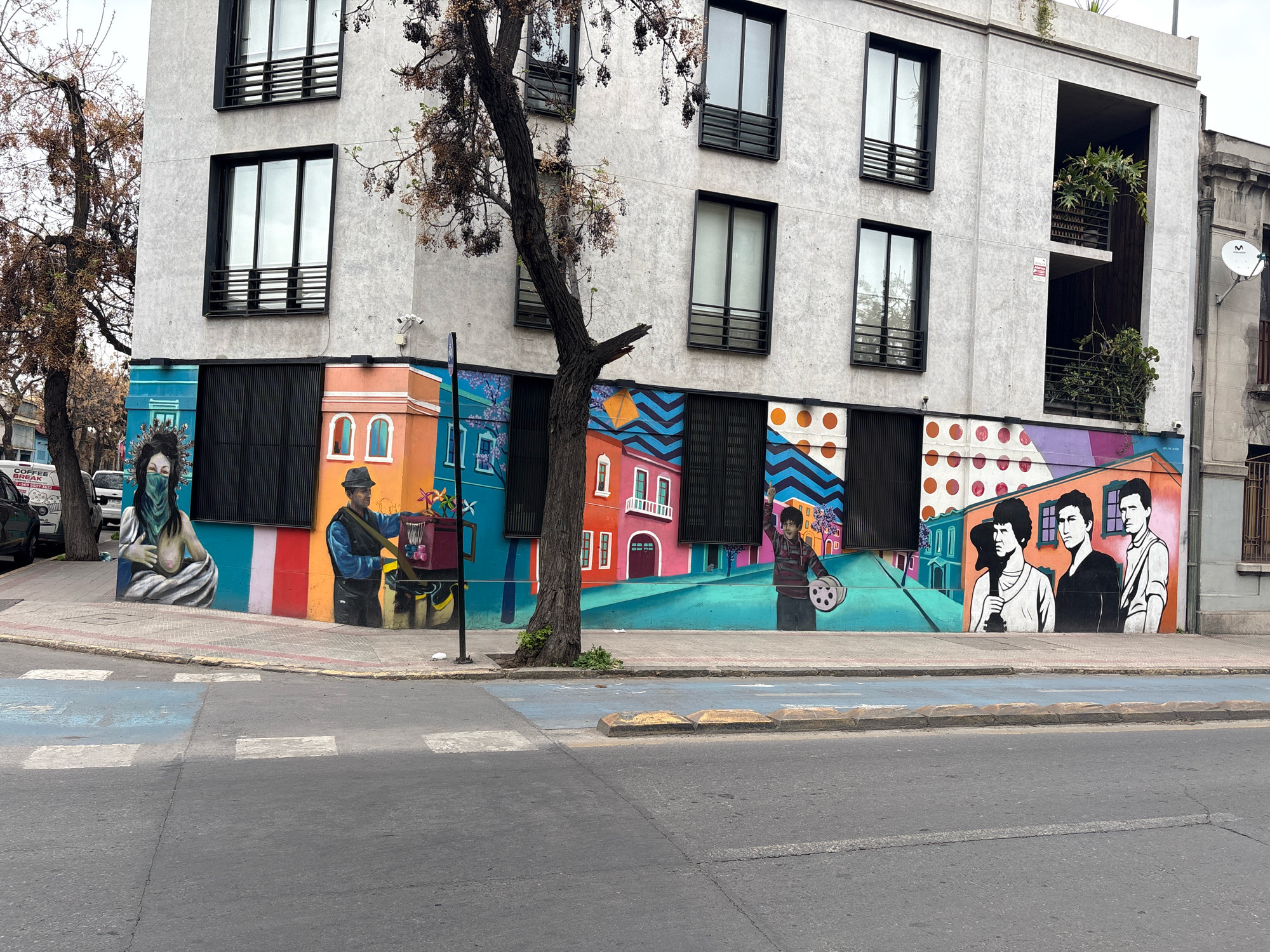 The image depicts a street scene in Santiago, Chile, featuring a modern building with a vibrant mural painted on its lower facade. The building itself is a multi-story structure with a light-colored, textured exterior. Several windows with dark frames are visible on the upper floors. A prominent tree with sparse, bare branches grows in front of the building, partially obscuring some of the mural.
The mural is highly colorful and depicts various figures and scenes. On the left side, a woman with a patterned headscarf and a green face covering is shown. Adjacent to her, a man wearing a hat and dark clothing is depicted with a cart filled with colorful items, including pinwheels. Further to the right, a young person in a striped shirt holds a film reel. The mural also features stylized buildings with colorful facades, geometric patterns, and a group of three men with distinct hairstyles. The artwork spans across multiple sections of the building's lower level, creating a dynamic and engaging visual.
The foreground of the image shows a street with a crosswalk marked by blue and white paint. A sidewalk with a raised curb separates the street from the building. The overall environment suggests an urban setting, with the cloudy sky indicating overcast weather. The presence of street markings and the building's architecture are consistent with a city environment like Santiago.