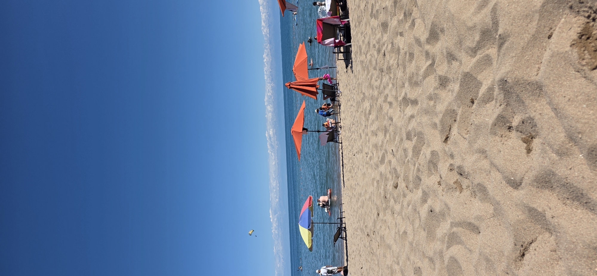 This is a photograph of a beach on a sunny day, likely in Bulan-Segettu, Kyrgyzstan, given the clear blue sky, calm water, and distant snow-capped mountains. The foreground is dominated by a sandy beach with footprints and textured sand. Several beach umbrellas are set up: one is multi-colored (yellow, red, blue), and the others are bright orange. Beach chairs are placed under some of the umbrellas.
In the water, several people are visible. Some are swimming or wading, while others are on inflatables. One person is on a paddleboard. In the distance, there are more people in the water and on the shore. A small yellow and black parasail is visible in the sky. The water is a deep blue, and the horizon is marked by a line of fluffy white clouds above the distant mountains. The overall atmosphere is one of leisure and recreation.