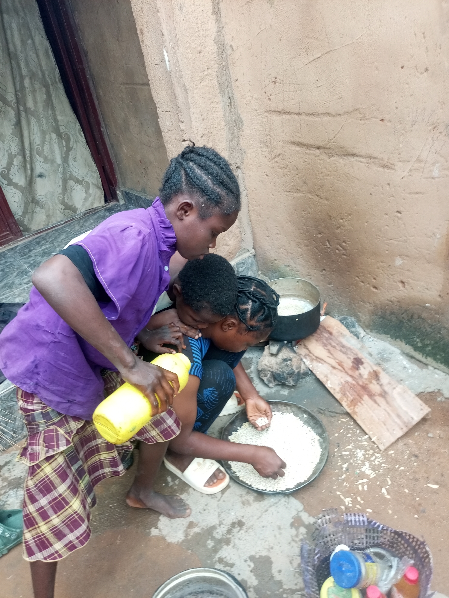 In Tempure, Nigeria, two young individuals are engaged in food preparation outdoors. The scene is rustic, with a rough, textured wall and a tiled floor. In the foreground, a person wearing a purple tunic and a plaid skirt is holding a bright yellow container, pouring its contents. Another person, to their right, is leaning over a metal bowl filled with white grains, sorting them with their hands. A pot of what appears to be food is simmering on a makeshift stove nearby, and a wooden plank rests on the ground. A woven basket containing various bottles and containers is also visible. The overall impression is one of everyday life and resourcefulness.