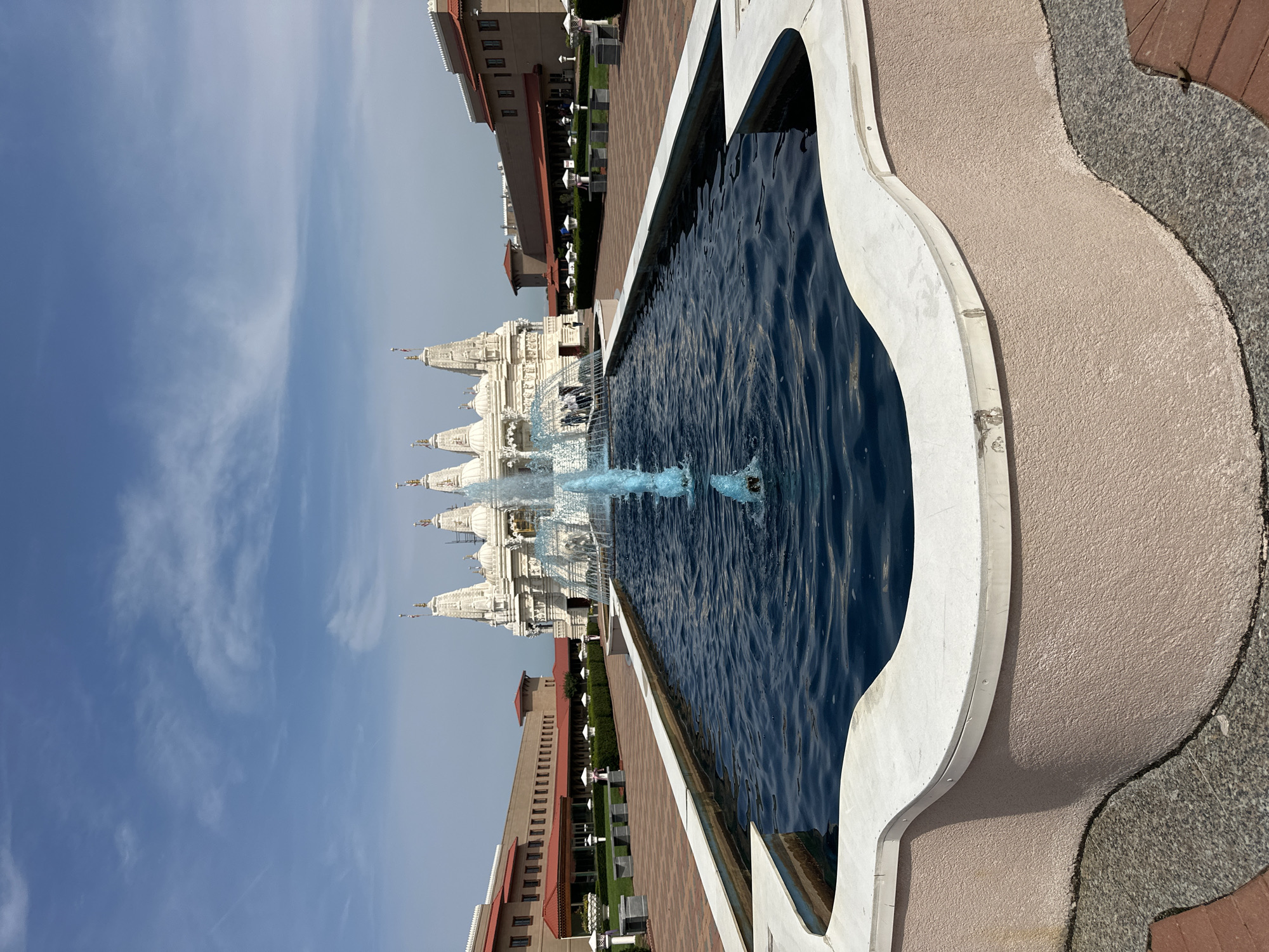 A formal garden path of brick and stone leads to a large white temple structure with multiple tiered spires. In the foreground is a large, dark blue rectangular pool with a decorative wavy edge made of white stone and a pink textured base. A fountain in the center of the pool shoots bright blue water upwards. The temple is set back from the pool, with a manicured lawn and hedges between them. Two long, low buildings with red tiled roofs flank the temple. The sky is blue with scattered white clouds.