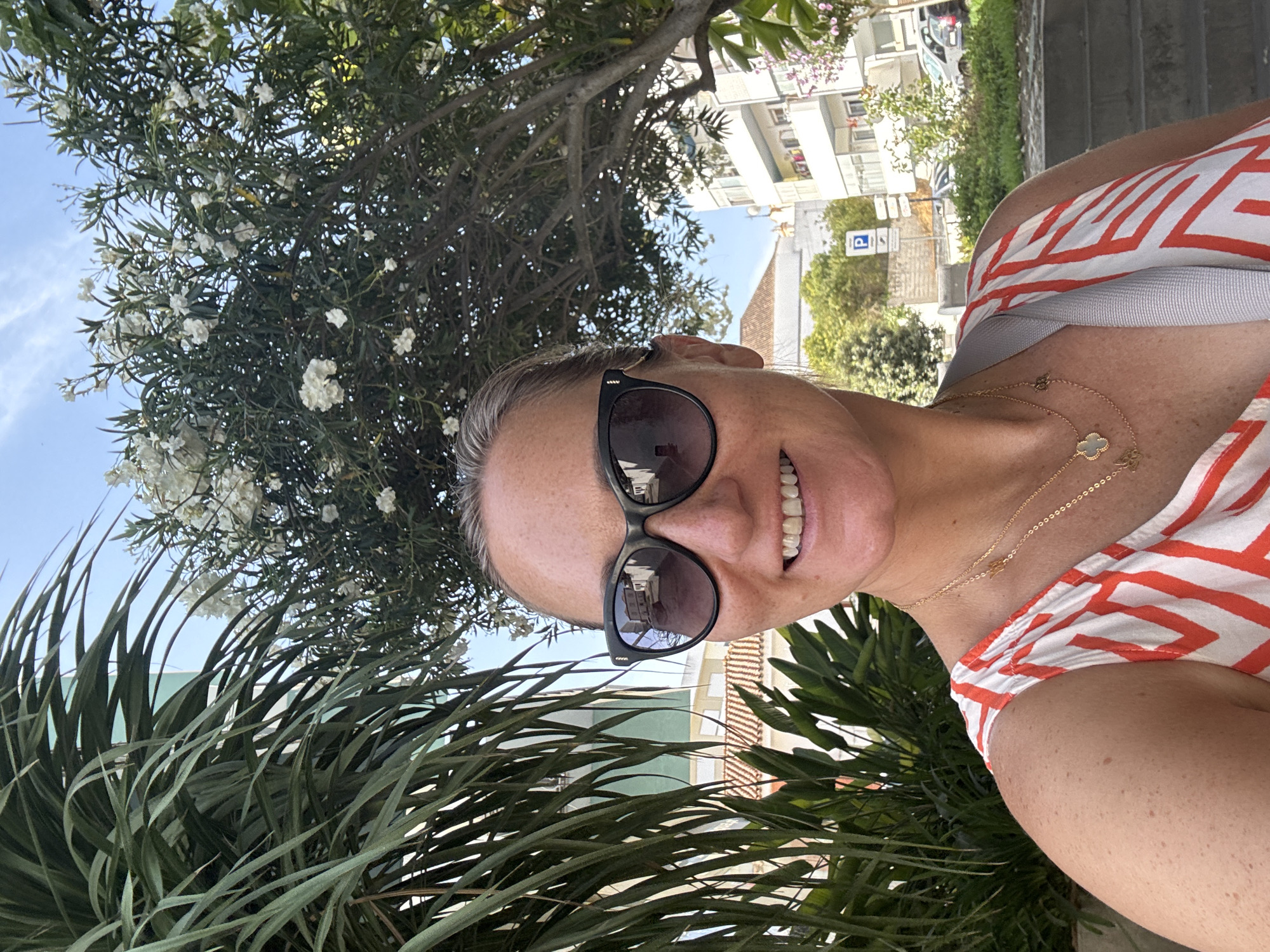 A mid-shot selfie of a woman with a smile is taken outdoors in a sunny environment. She is wearing large, dark sunglasses and a white and orange striped top. Her hair is pulled back, and she has freckles on her face and arms. She is wearing two gold necklaces, one with a clover-shaped pendant and another with a butterfly pendant. Behind her, there are palm fronds on the left, a tree with white flowers in the center, and buildings with balconies and parked cars in the background. The location appears to be a street in Tavira, Portugal, as indicated by a parking sign with the letter "P" in the background.