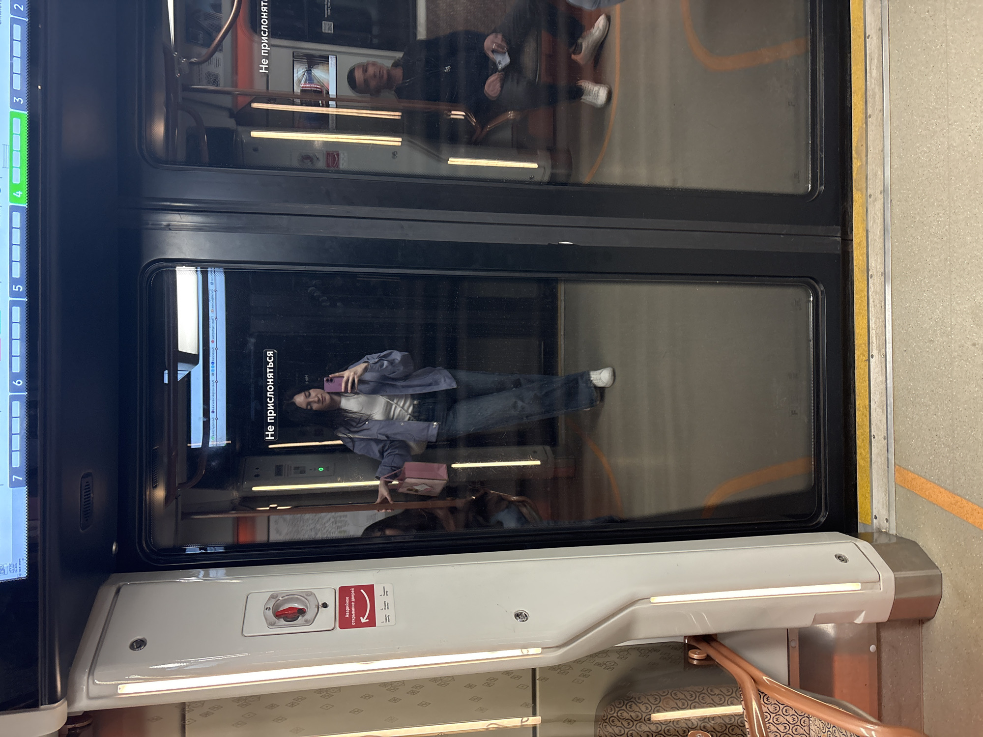 The image shows the interior of a Moscow Metro train. Visible are two passengers reflected in the window. The door features signage in Russian, including "Не прислоняться" (Do not lean) and emergency door release instructions. The train's digital display indicates stops numbered "3, 4, 5, 6, 7". A partial view of a bag suggests a passenger's personal belongings. The scene is brightly lit by overhead lighting. The Moscow city location is indicated by the standard train equipment, signage, and interior design typical of the Moscow Metro.