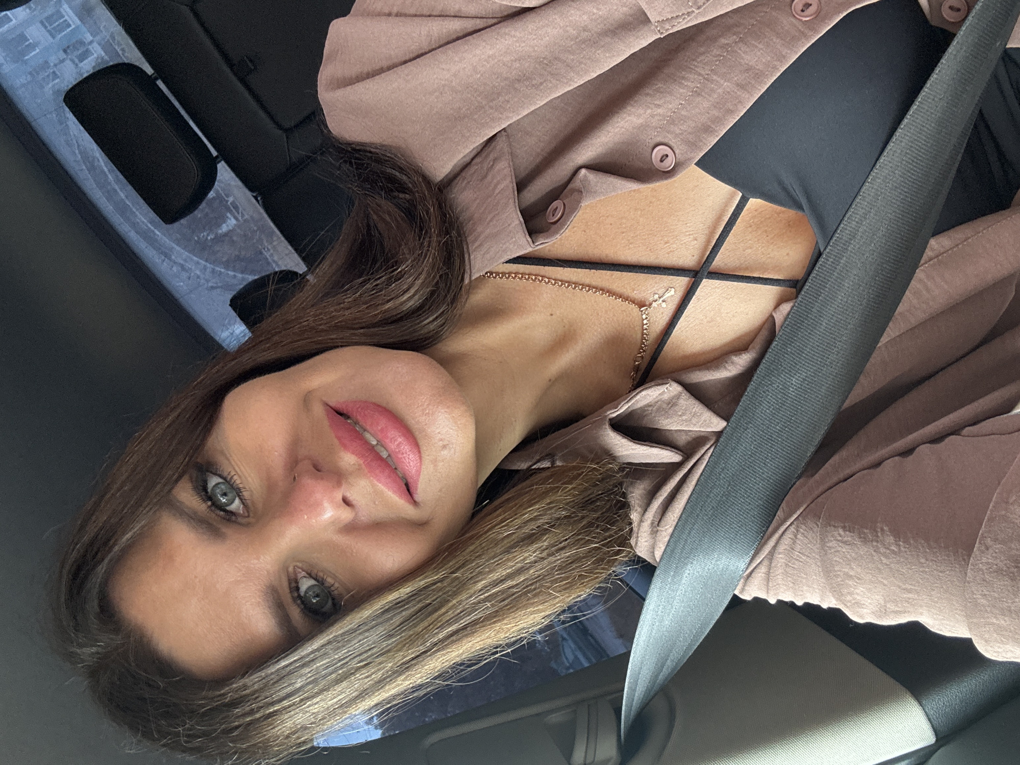 A close-up, waist-up selfie of a fair-skinned woman with brown hair and blue eyes is captured inside a vehicle. She is wearing a pink button-down shirt over a black camisole and a gold cross necklace. A gray seatbelt is diagonally positioned across her chest. The background shows the interior of a car, including a headrest and what appears to be a window with a view of buildings outside. The image is set in Chaikovsky, Russia.