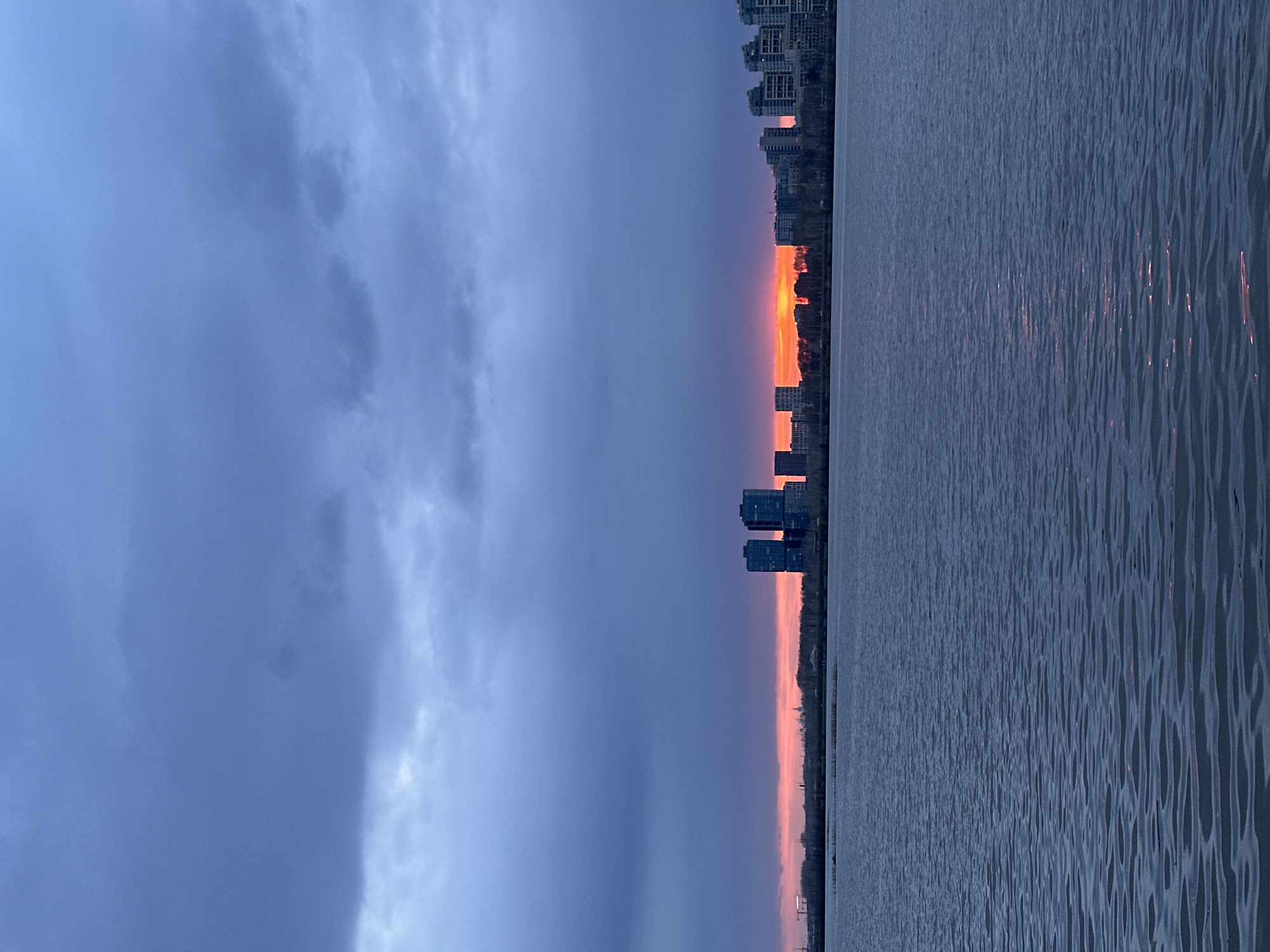 The image shows a body of water, possibly the Kazanka River, with buildings in the distance likely located in Kazan, Russia. The sky is predominantly blue with dark clouds on the left side. A vibrant sunset casts a reddish-orange glow on the horizon between the water and buildings, and reflects on the surface of the water. The water is calm with small ripples. The buildings appear to be a mix of high-rise residential and commercial structures.