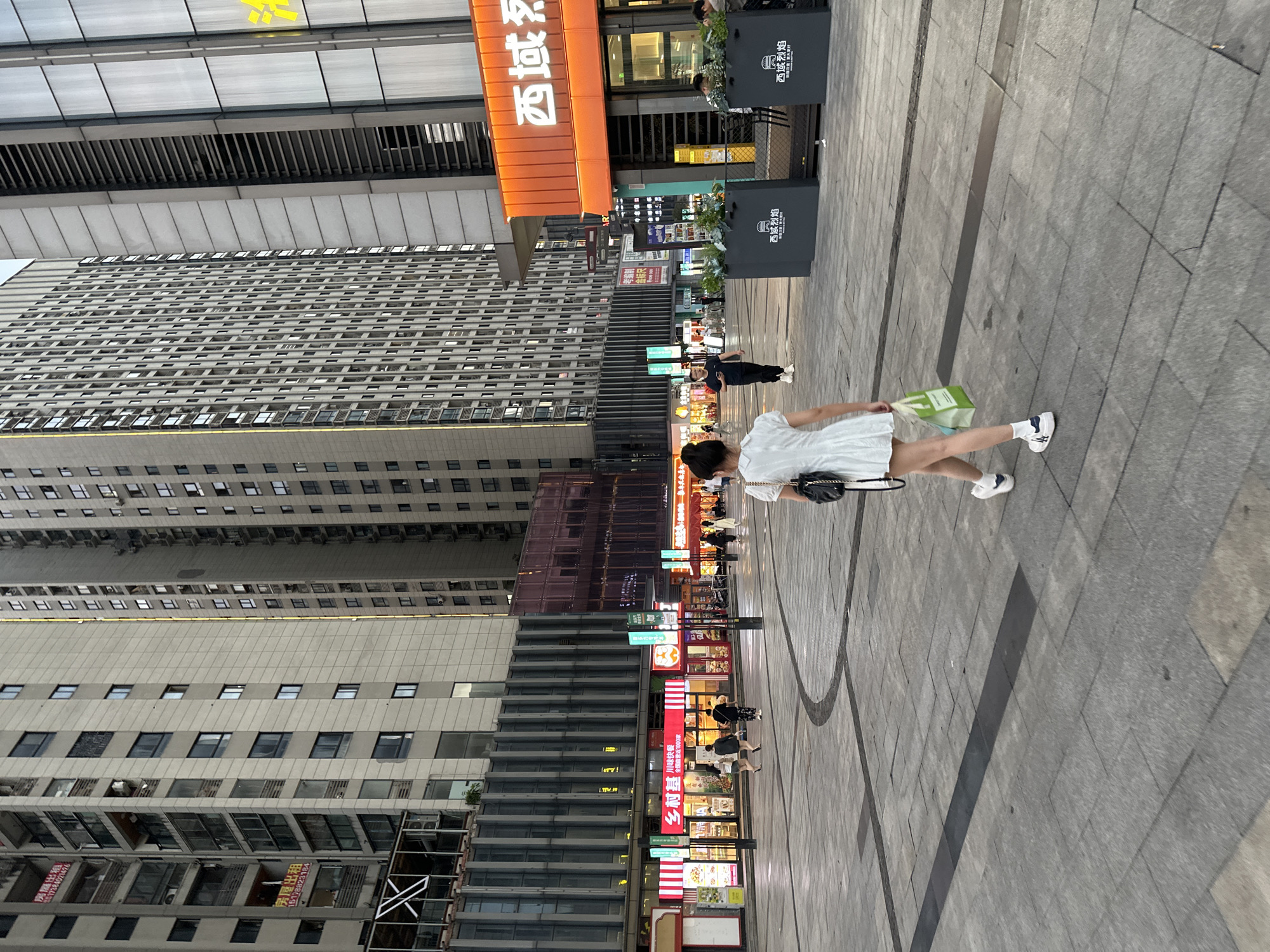 A woman in a white dress walks on a gray brick sidewalk in the Chongqing High-tech Zone, China. She carries a green shopping bag. The environment includes tall buildings, including one with a sign that reads "Xi He Yi Duo." A number of storefronts are visible. Various signs with Chinese writing are present. The sidewalk also has circular designs.