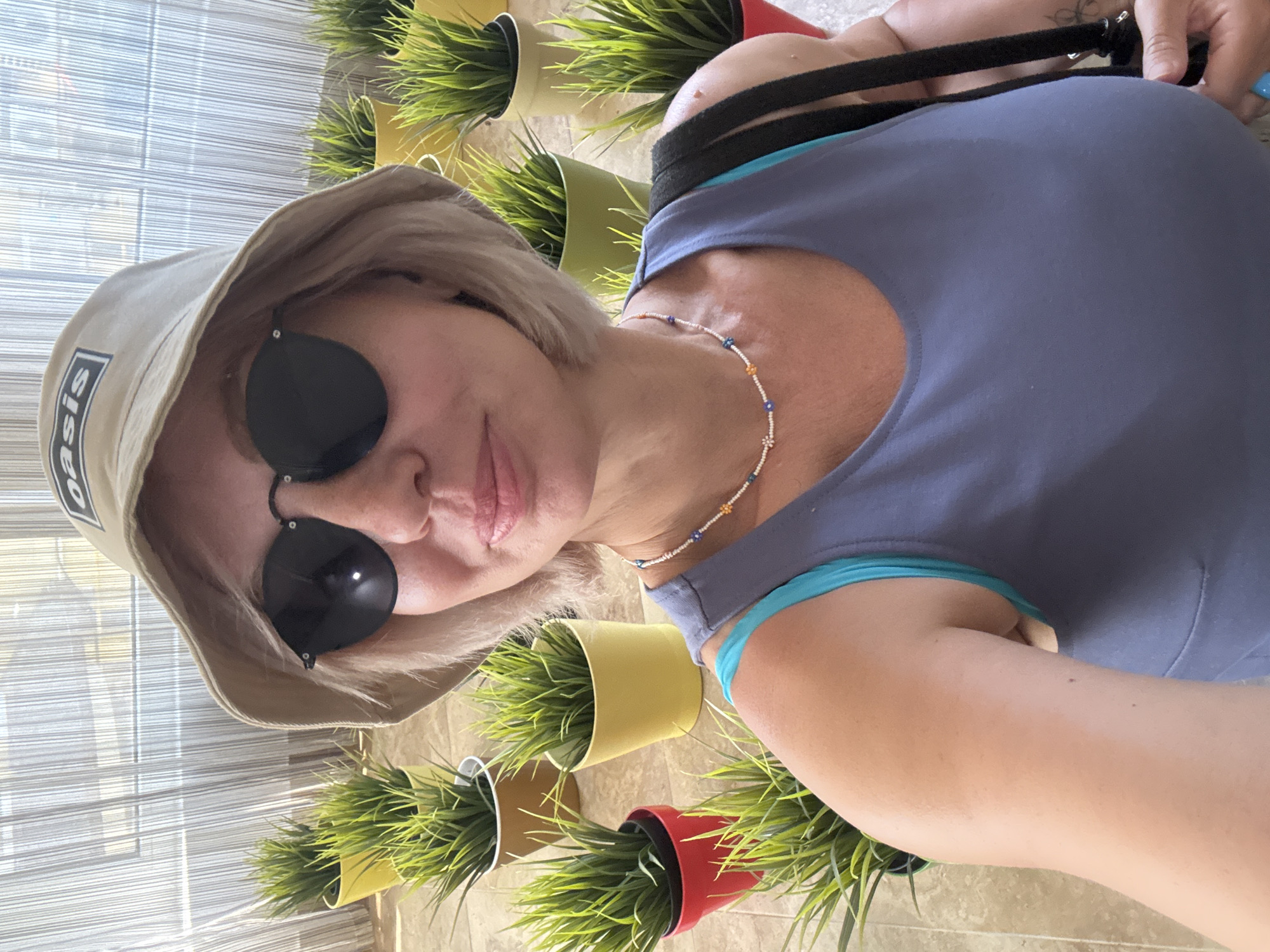 A woman with blonde hair, wearing a beige bucket hat with "Oasis" printed on it, black sunglasses, and a purple tank top over a turquoise bra, takes a selfie. She is also wearing a beaded necklace with colorful floral designs. The background features several potted artificial plants arranged on a tiled floor, with sheer white curtains in the background. The scene appears to be indoors, possibly in a hotel or resort, and the lighting suggests daytime. There is no specific indication of the location being within the city of Sirius, Russia.