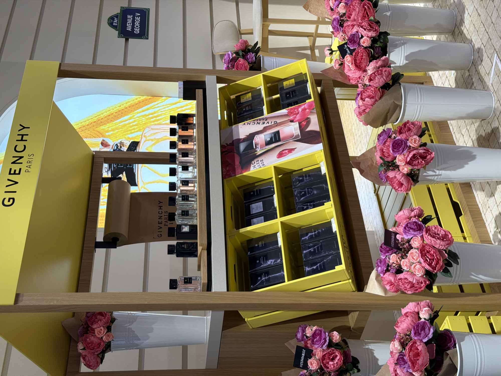 The image depicts a retail display featuring Givenchy perfumes. The display is brightly lit with a yellow overhead sign bearing the "GIVENCHY PARIS" logo. Various bottles of perfume are arranged on shelves, with some displayed prominently on a wooden shelf in the upper section and others in yellow bins below. The lower shelves are filled with boxed perfume sets. Decorative elements, including bouquets of pink and purple artificial flowers in white vases, are strategically placed around the display, both on higher shelves and at the bottom. In the background, a sign on a wall reads "8° Arr. AVENUE GEORGE V," suggesting a Paris street address. The overall environment appears to be an indoor retail space, likely a department store or a dedicated brand boutique, with a light-colored tiled floor and white paneled walls. There are no people visible in the image. The scene is clean and well-organized, highlighting the products and the brand's aesthetic. The location is not Arnavutköy, Turkey; the sign clearly indicates an address in Paris, France.