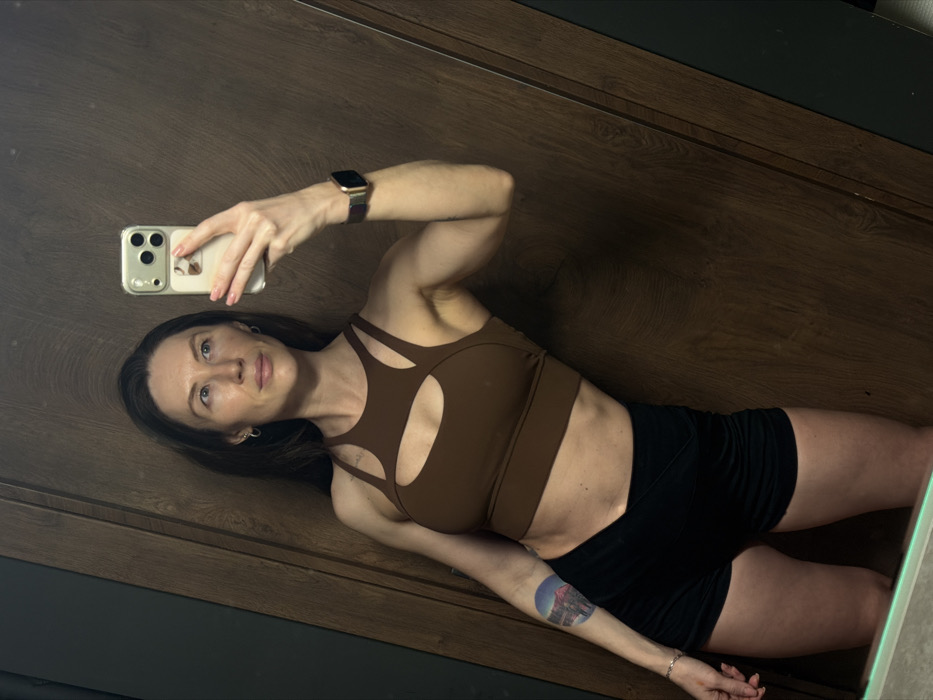 A fair-skinned woman takes a selfie in a mirror. She wears a brown cut-out sports bra and black shorts. Her dark hair is down, and she has a watch on her left wrist. A tattoo is visible on her left arm. The background shows a wood-paneled wall with a darker trim at the top. The phone in her hand is gold.