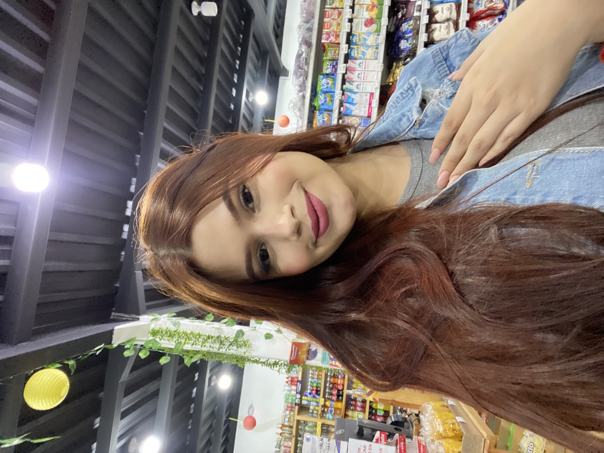 A young woman with long, wavy, reddish-brown hair, dark eyes, and pink lipstick is captured in a selfie-style portrait. She wears a grey top beneath a light blue denim jacket, with her left hand, adorned with light pink manicured nails, resting on her chest. She displays a soft smile, looking directly at the camera.
The setting is an indoor commercial space, likely a supermarket or convenience store. The background reveals multiple shelves stocked with various packaged products, including beverages and snacks. The ceiling features exposed dark metal beams with bright, circular light fixtures. Decorative elements include artificial green vines draped across overhead structures and two spherical lanterns, one yellow and one orange-red.
The activity is the woman taking a self-portrait within this retail environment. The mood appears casual and pleasant.
Visible text is scarce due to blurriness and perspective. Numbers "02.40" and "99.00" are visible on white shelf labels in the background. Partial product names or descriptions like "Coco," "tro," and "ate" (potentially from "Colgate") can be vaguely discerned on some packaging on the shelves. The general store environment is consistent with commercial settings found in places such as Juan Griego, Venezuela.