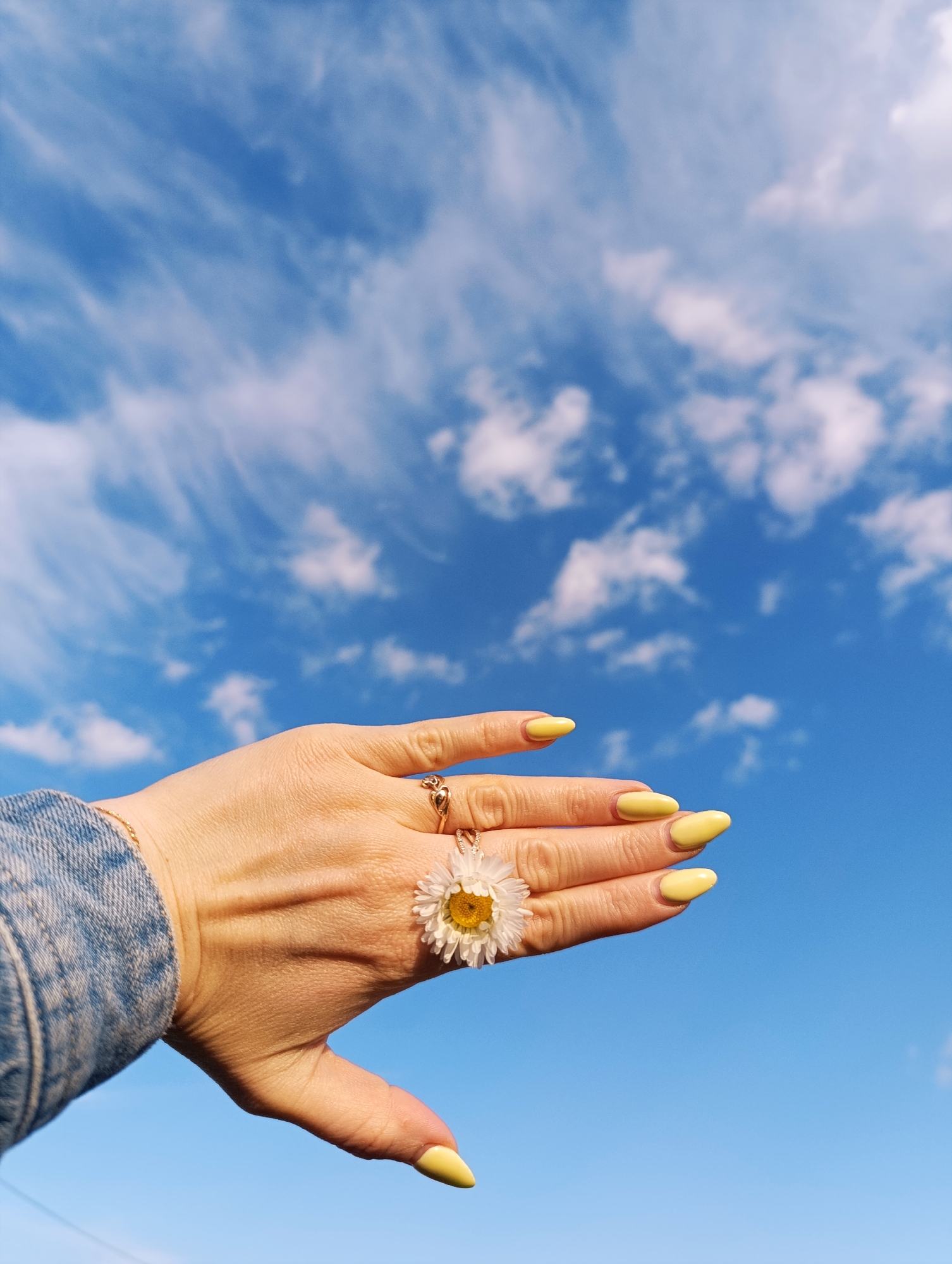 A close-up, upward-angled shot captures a human hand, likely female, held against a vibrant blue sky. The hand features bright yellow, oval-shaped manicured nails. A gold-toned ring with an intricate, possibly heart-shaped, design is visible on the ring finger. A small white daisy flower, with a prominent yellow center, is tucked between the index and middle fingers, giving the appearance of a natural ring. A thin, gold-toned chain bracelet adorns the wrist, which is partially covered by a blue denim sleeve, suggesting a jacket or shirt.
The background is a clear, bright blue sky filled with scattered white clouds. These include wispy cirrus clouds stretching across the upper part of the frame and some denser, fluffy cumulus clouds. The lighting indicates bright daylight, likely midday or afternoon, under fair weather conditions. The scene evokes feelings of nature, freshness, and the outdoors, possibly during spring or summer. The specific location is unknown. There is no visible text in the image.