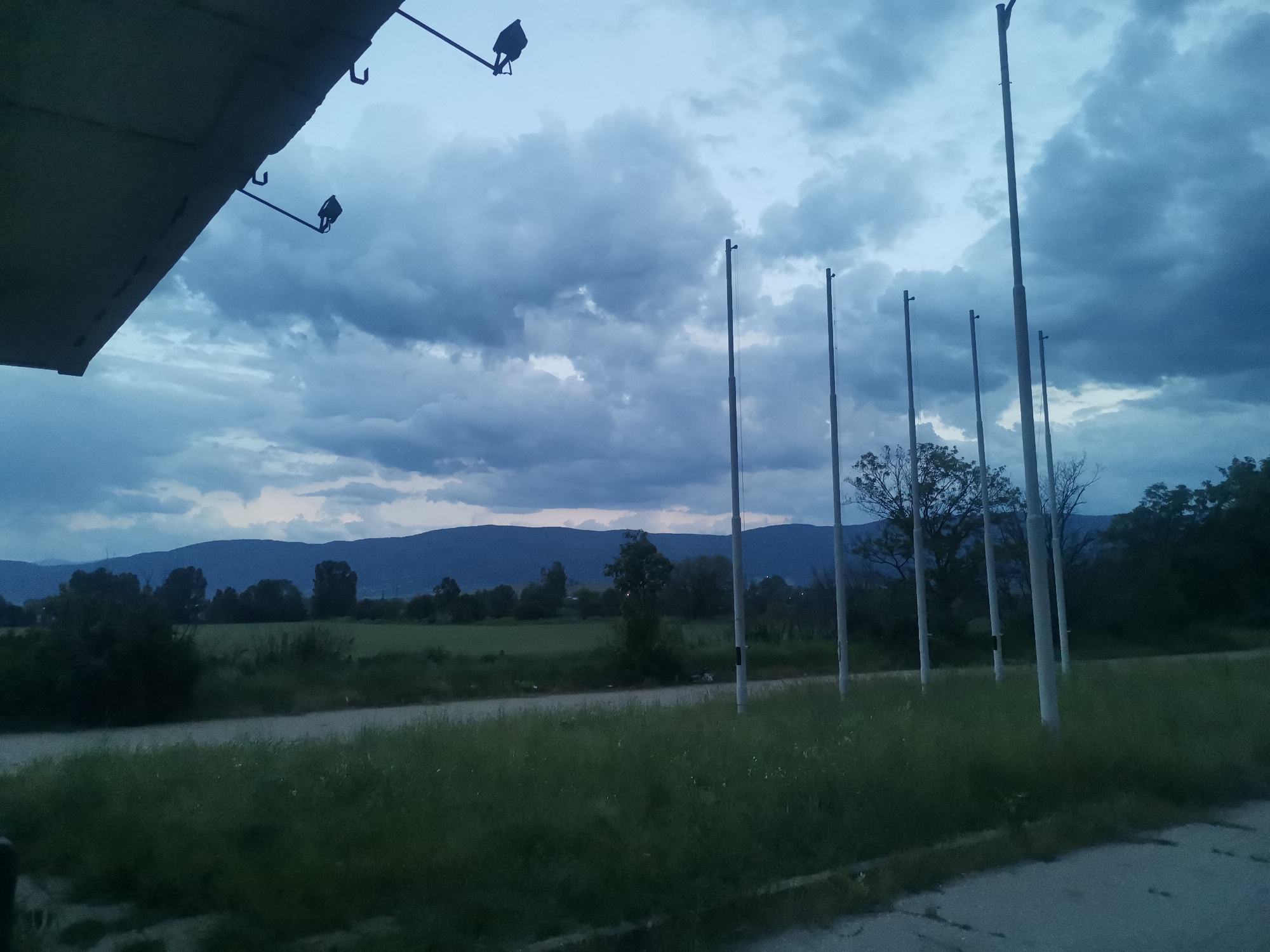 The image presents an outdoor, rural landscape in Zhivkovo, Bulgaria, under a heavily overcast sky, suggesting either dusk, dawn, or a deeply clouded day. In the upper left corner, the underside of a large, flat structure, possibly an awning or roof overhang, is visible, featuring several metal hooks and two small, dark light fixtures attached to thin rods.
Stretching across the mid-ground from right to left is a line of at least six tall, slender poles, which could be flagpoles or light posts. These poles are set within a grassy field, through which a dirt or gravel path or road runs horizontally. Further back in the mid-ground, scattered trees dot the landscape beyond the path. The background is dominated by a range of dark, undulating hills or mountains, silhouetted against the dramatic, cloudy sky. The clouds are dense and varied in tone, ranging from lighter greys to deep blues and purples.
The scene is tranquil and devoid of human presence or activity. The subdued lighting and dark clouds create a somewhat somber or contemplative mood. There is no visible text in the image.