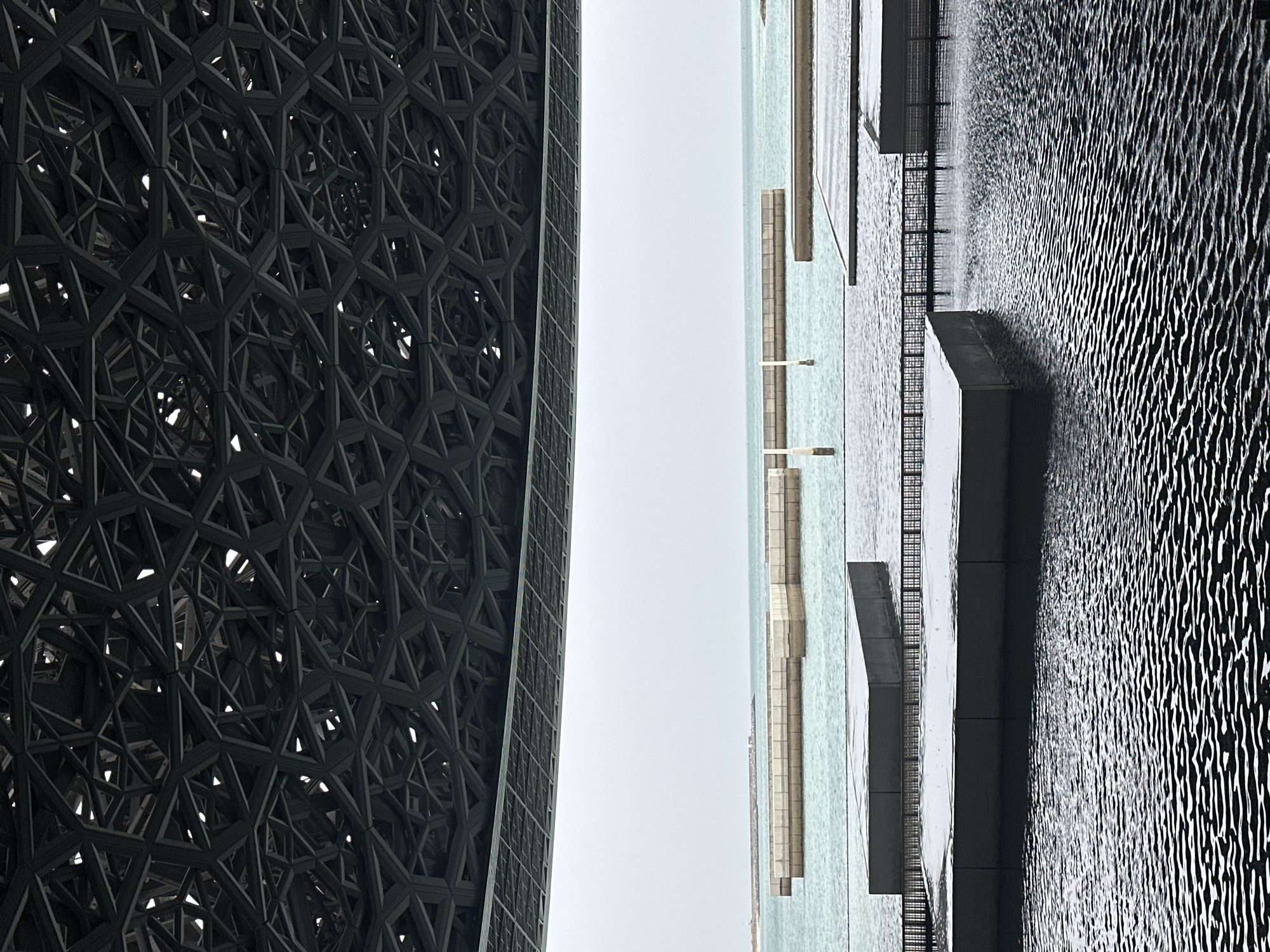 The image shows a section of the Louvre Abu Dhabi in Abu Dhabi, United Arab Emirates. On the left is a portion of the museum's dome, which features an intricate, geometric pattern of overlapping metal structures. To the right of the dome is a light blue expanse of water that appears to be the sea, with several elevated concrete structures and walkways extending over it. The water surface shows distinctive wave patterns, creating a textured effect. The sky is a uniform pale gray, suggesting overcast conditions. There is no indication of people or interaction within the frame.