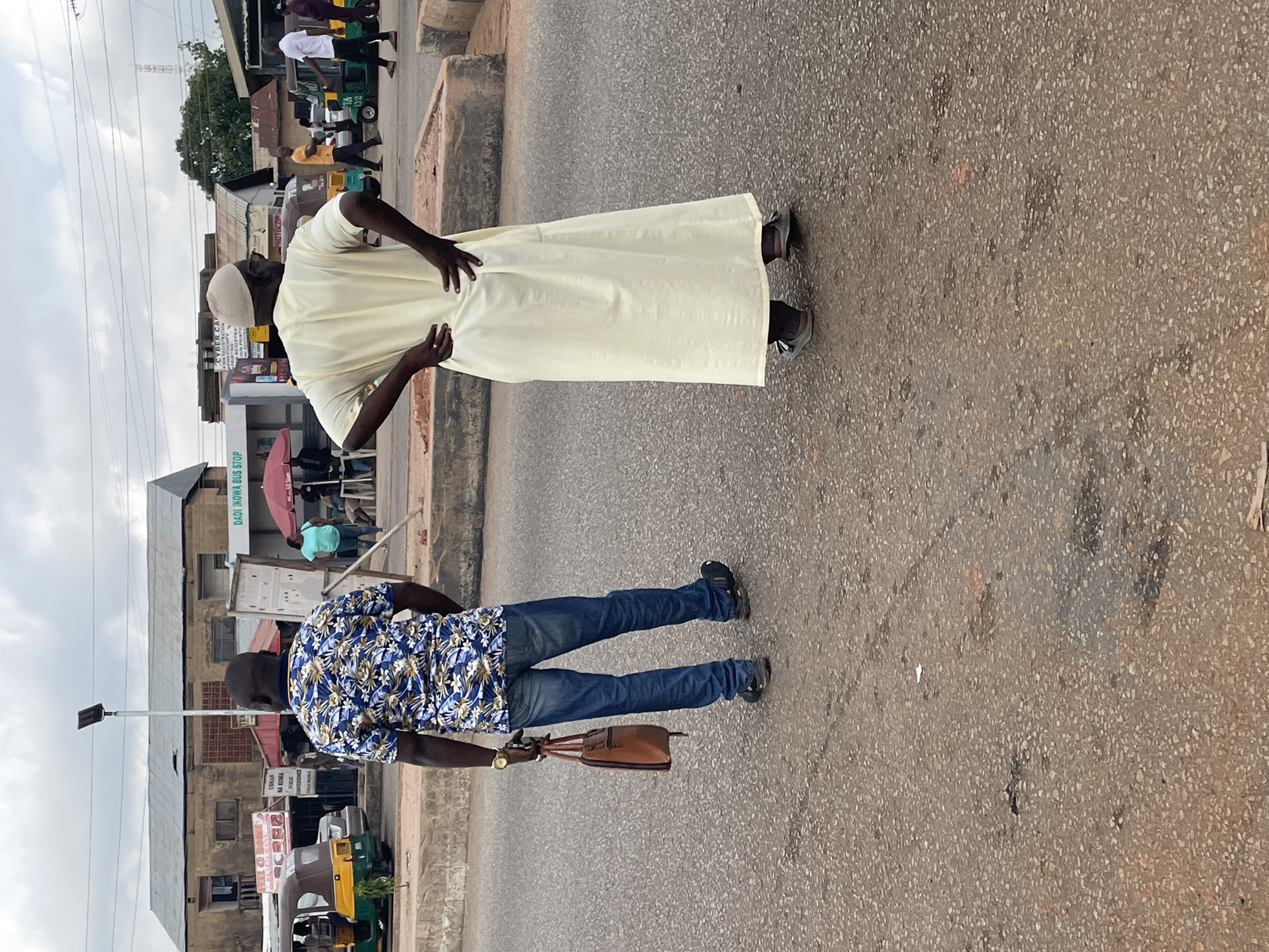 The image captures a street scene in Gura Top, Nigeria, with a focus on two men standing on a paved road. The man on the left, facing away from the camera, wears a blue and white floral short-sleeved shirt, blue jeans, and dark sneakers. He carries a brown leather bag in his right hand and has a gold watch on his left wrist. The man on the right, also facing away, is dressed in a long, light yellow robe and a white cap. He has his hands on his hips. In the background, a bus stop labeled "DADI INIOWA BUS STOP" is visible, along with several buildings and vehicles, including auto-rickshaws. The sky is overcast.